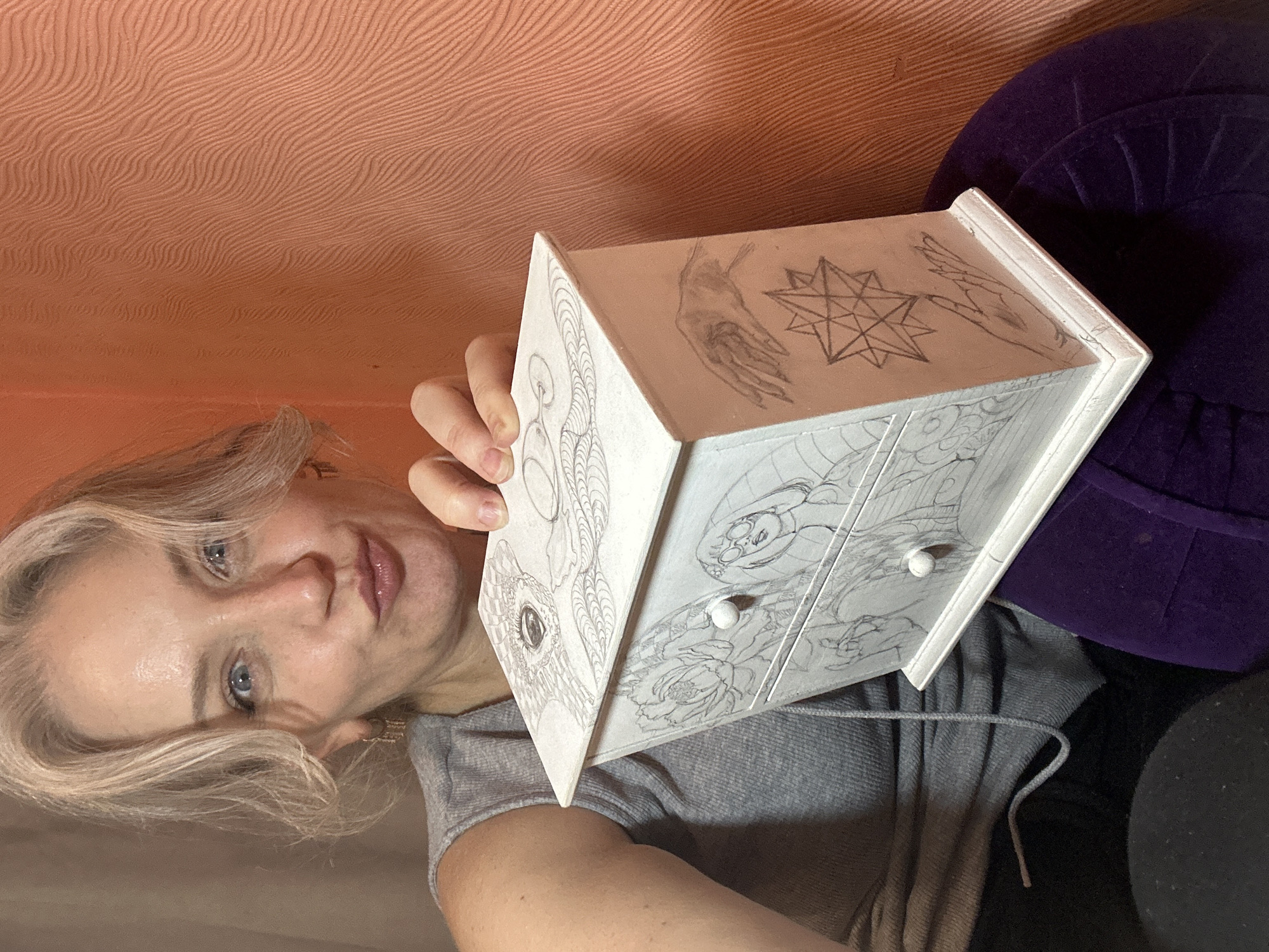 A woman with blonde hair and blue eyes is holding a white jewelry box decorated with pencil drawings. The box has two drawers and is adorned with various sketches, including an eye on the lid, flowers, a person wearing glasses, hands, and a star. The woman is wearing a grey tank top and is positioned indoors against a peach-colored textured wall. The scene appears to be a personal setting, potentially a home. There is no information in the image to suggest a specific location in the city of Kyiv, Ukraine.