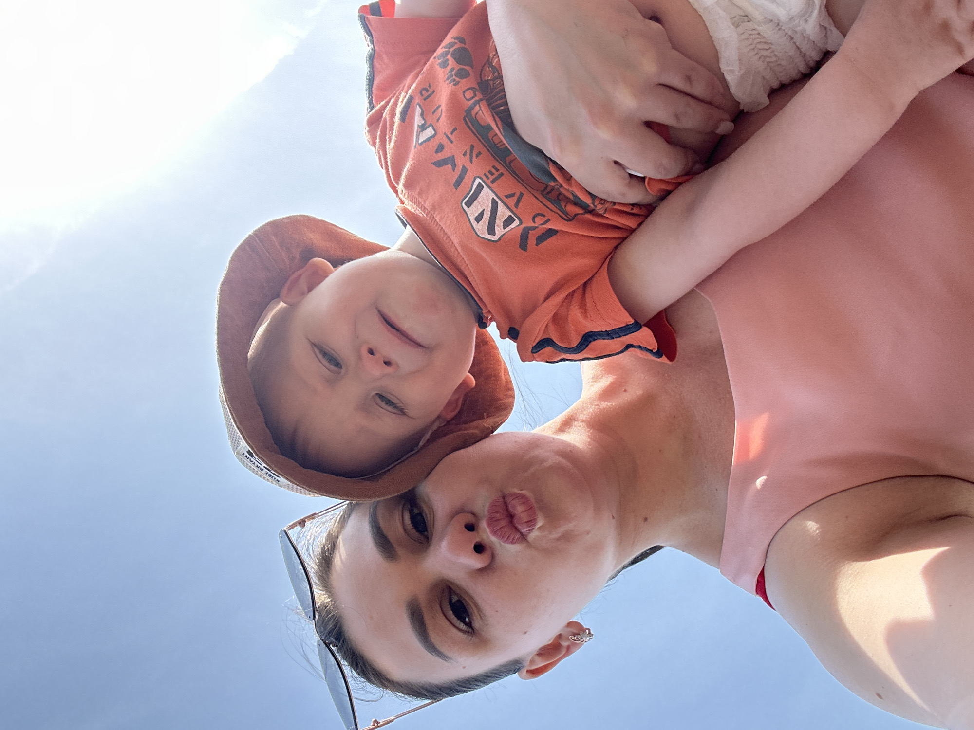 A woman and a young child are in the foreground of a low-angle shot taken outdoors, likely in Chaikovsky, Russia, based on the prompt's instruction. The woman is holding the child close; the child is partially obscuring the woman's upper chest and shoulder. The woman is looking directly at the camera with pursed lips, wearing sunglasses perched on her head. The child is also looking at the camera. The child is wearing an orange t-shirt with writing and graphics and a brown sun hat. The background consists of a clear, light blue sky.