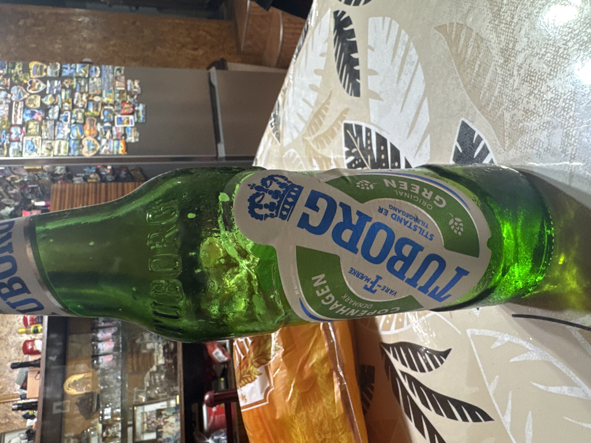 The image captures a close-up, slightly tilted view of a green Tuborg beer bottle resting on a table. The bottle is visibly cold, covered in condensation. Its label features the prominent blue text "TUBORG" with a crown logo above it. Other visible text on the label includes "ORIGINAL", "GREEN", "Copenhagen", "DENMARK", "VARE-MÆRKE", and "STILSTAND ER TILGÆNGANG".
The table is covered with a light-colored tablecloth patterned with dark, stylized leaves. In the background, to the left, a refrigerator or metal surface is visible, adorned with numerous small, colorful magnets. Below the bottle, there's a blurry orange object, possibly a plastic bag. Reflections of various objects, including shelves and other bottles, are visible in the background, suggesting an indoor setting, likely a kitchen or dining area in Sarnevo, Bulgaria. The lighting appears artificial, indicating an indoor scene without specific time-of-day cues. No people are present, and no particular activity is depicted beyond the bottle resting on the table.