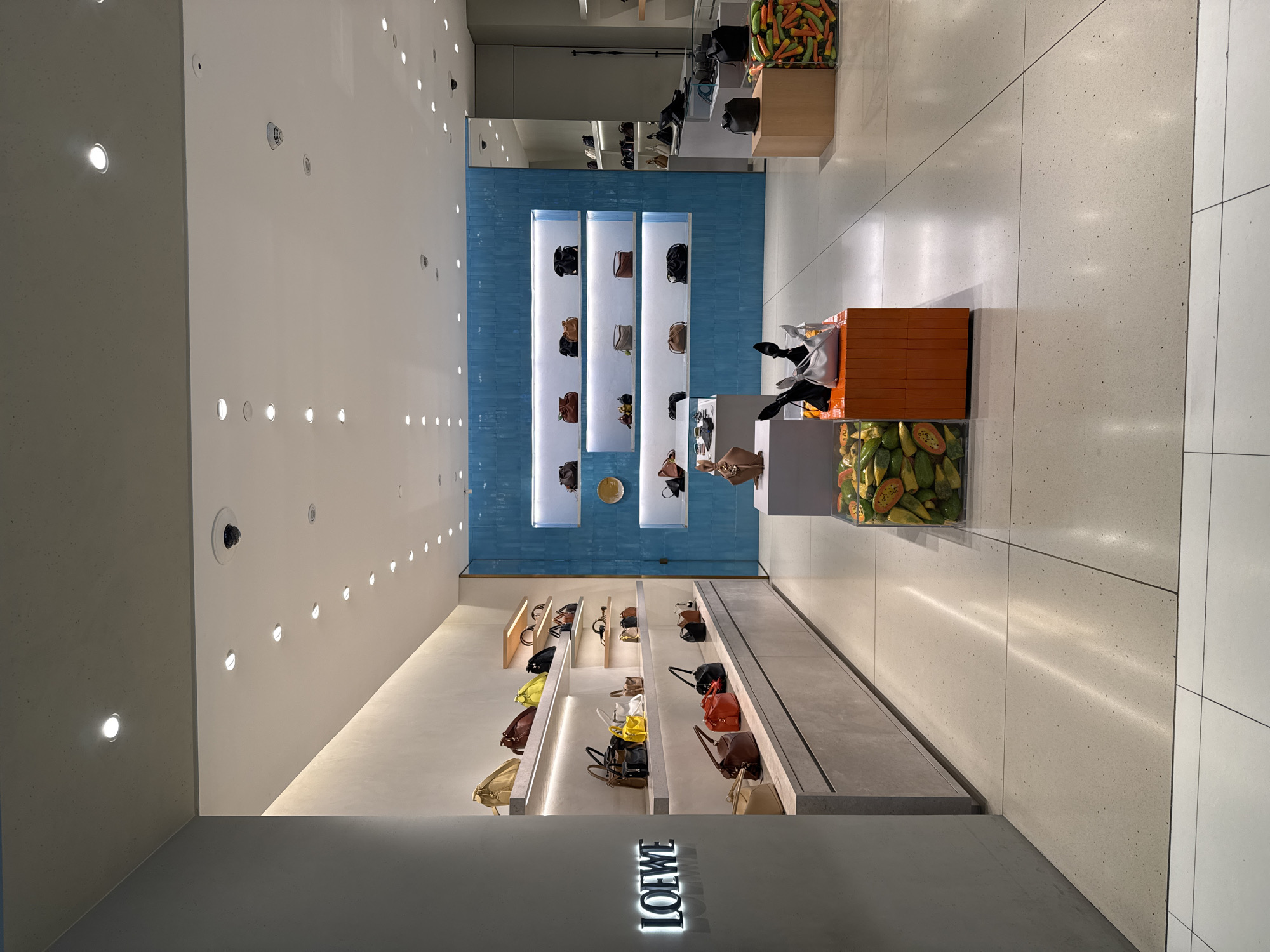 The image shows the interior of a Loewe boutique, likely located in a shopping mall in the city of Nong Prue Subdistrict, Thailand. The boutique features a minimalist design with white and gray walls, polished concrete floors, and bright, focused lighting.
The main display area showcases a variety of Loewe handbags on illuminated shelves. The bags are arranged in rows, with different styles and colors visible, including tan, burgundy, yellow, black, and red. The shelves are recessed into a light-colored textured wall, with a subtle upward glow.
To the right of the handbag displays, a section of wall is covered in vibrant blue, textured tiles, creating a striking visual contrast. More handbags are displayed on shelves within this tiled section.
In the foreground, there are display cubes. One cube is orange and contains abstract black and silver objects that resemble origami birds or bows. Another cube is clear and filled with cut papayas and avocados, adding a touch of natural color and texture to the retail environment. A further display cube to the right contains what appears to be a collection of chili peppers in various shades of green and orange.
The "LOEWE" logo is prominently displayed in illuminated lettering on the left-hand wall. The overall impression is one of luxury and modern design, typical of high-end fashion retail spaces. There are no people visible in the image.