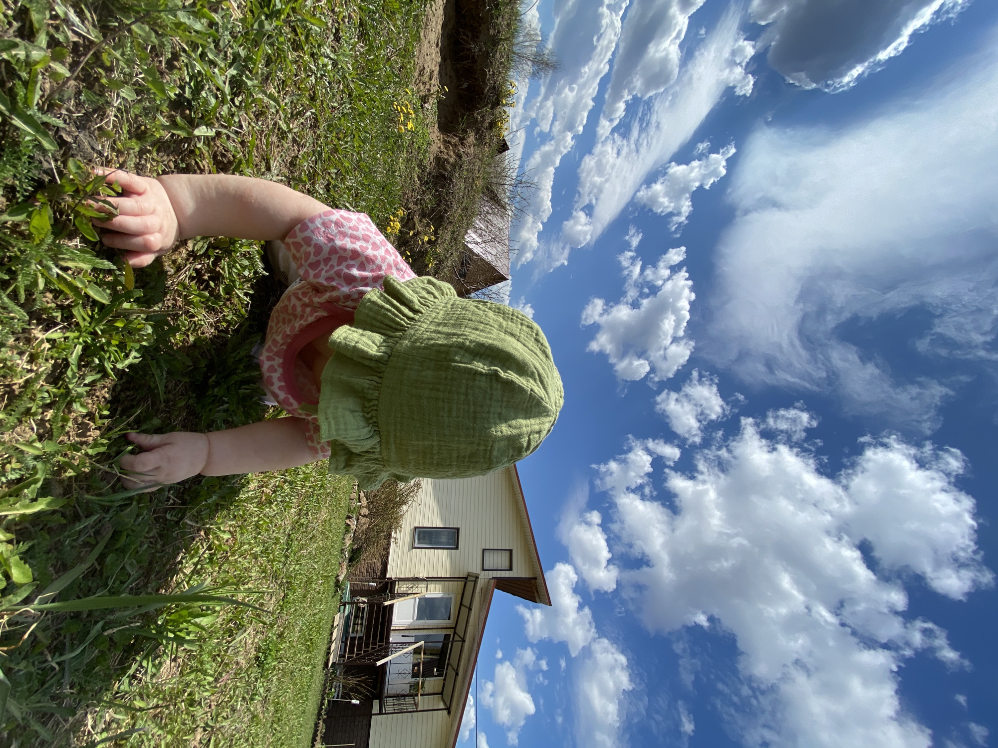 A baby wearing a green bonnet and a pink dress with hearts is exploring grass near a house. The house is yellow with a brown roof, a porch with railings, and windows. The sky above is blue with white clouds. The location is Фоки, Russia. The baby is reaching for plants in the grass.