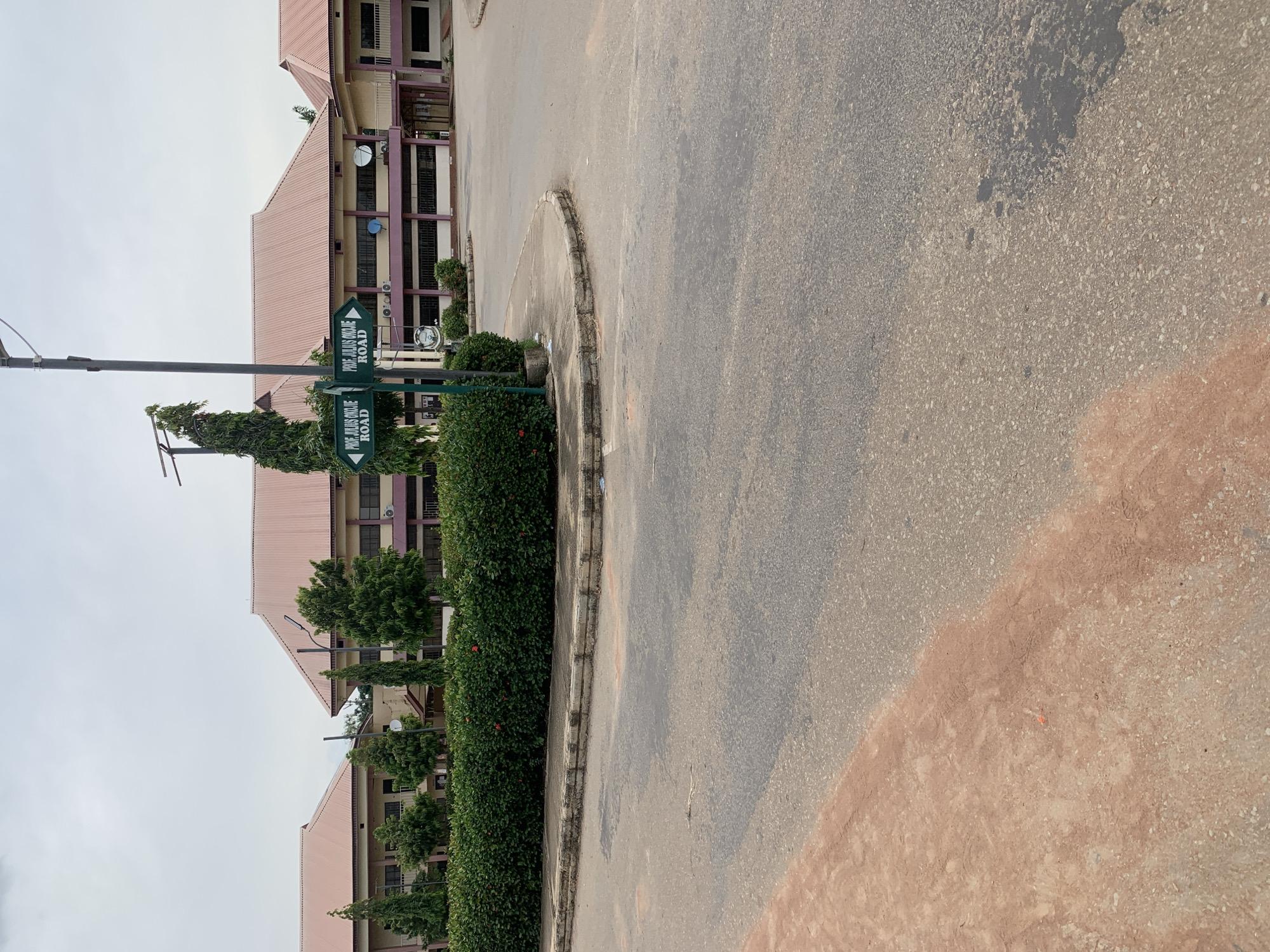 This media file depicts an outdoor scene in Wukari, Nigeria. The primary subjects are a two-story building with a pink-tiled roof and a paved road. The building features a beige facade with purple accents around the windows and has visible air conditioning units and satellite dishes on its exterior. Green, well-maintained bushes and trees line the front of the building.
A prominent object in the foreground is a street signpost with two green directional signs. One sign points left and reads "PROF. JULIUS OJOJE ROAD." Another sign, positioned below the first, also points left and contains the same text, "PROF. JULIUS OJOJE ROAD." A tall, slender lamp post is also visible on the left side of the frame.
The scene is set on an asphalt road that curves around a small, concrete-bordered island covered in greenery. The road appears to have some wear and tear, with patches of discoloration. The sky is overcast, suggesting a cloudy or potentially rainy day. The overall mood is neutral and observational. No people are visible in the frame, and no specific activity or event is taking place; it appears to be a static view of the location. The presence of the road sign indicates that this is a named street within the area.