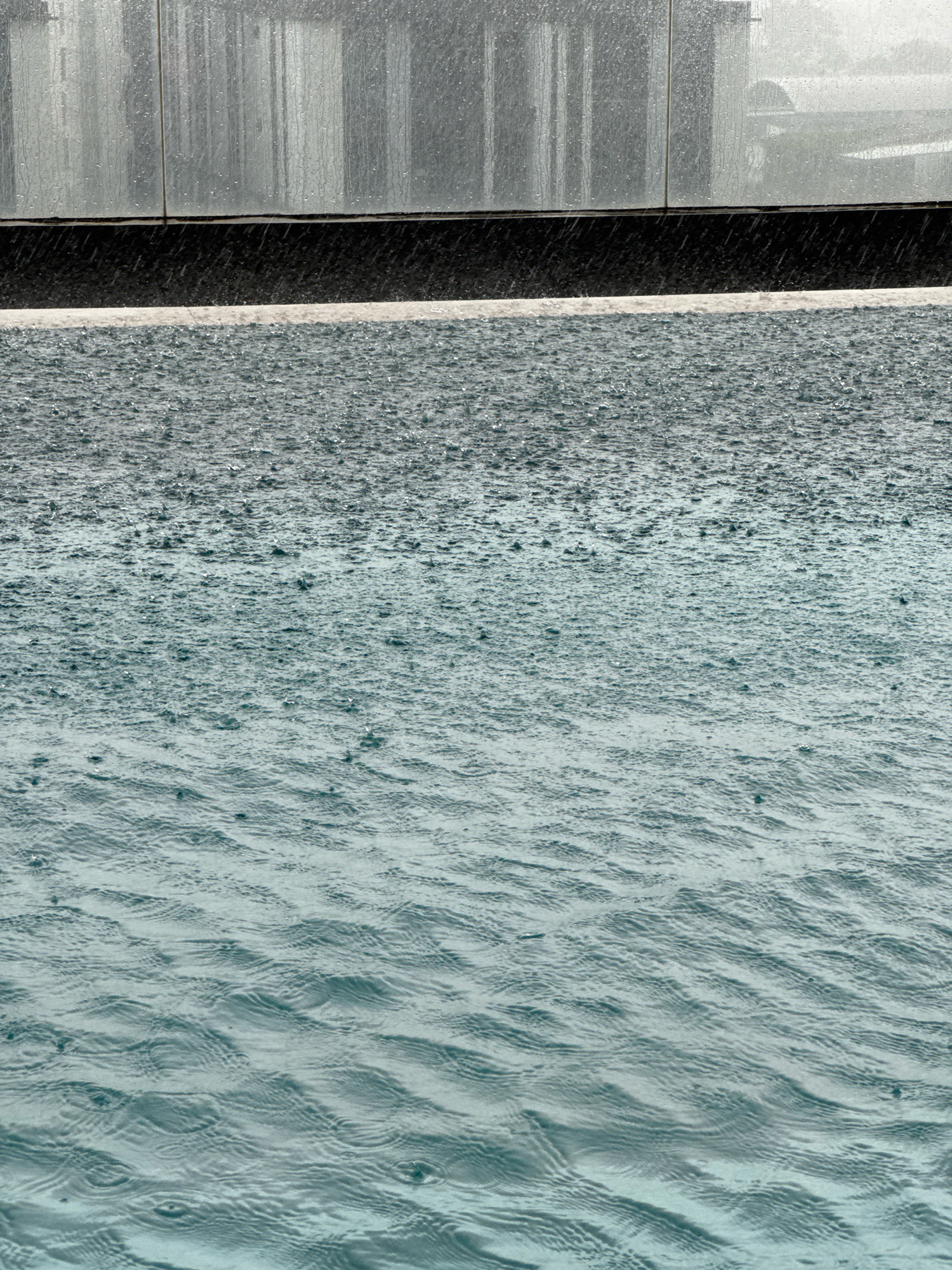 An exterior, medium shot captures a close-up view of heavy rainfall impacting a swimming pool. The water's surface is heavily disturbed by countless raindrops creating ripples and splashes. The pool edge is visible in the foreground, a light-colored coping separating it from a dark, textured band below. Beyond this, the facade of a modern building is seen, with a reflective, glass-like surface featuring vertical lines and water streaks. The scene is devoid of people. The environment suggests an urban setting, possibly a hotel or residential complex, within Samrong Nuea Subdistrict Municipality, Thailand, given the weather conditions and architectural style.