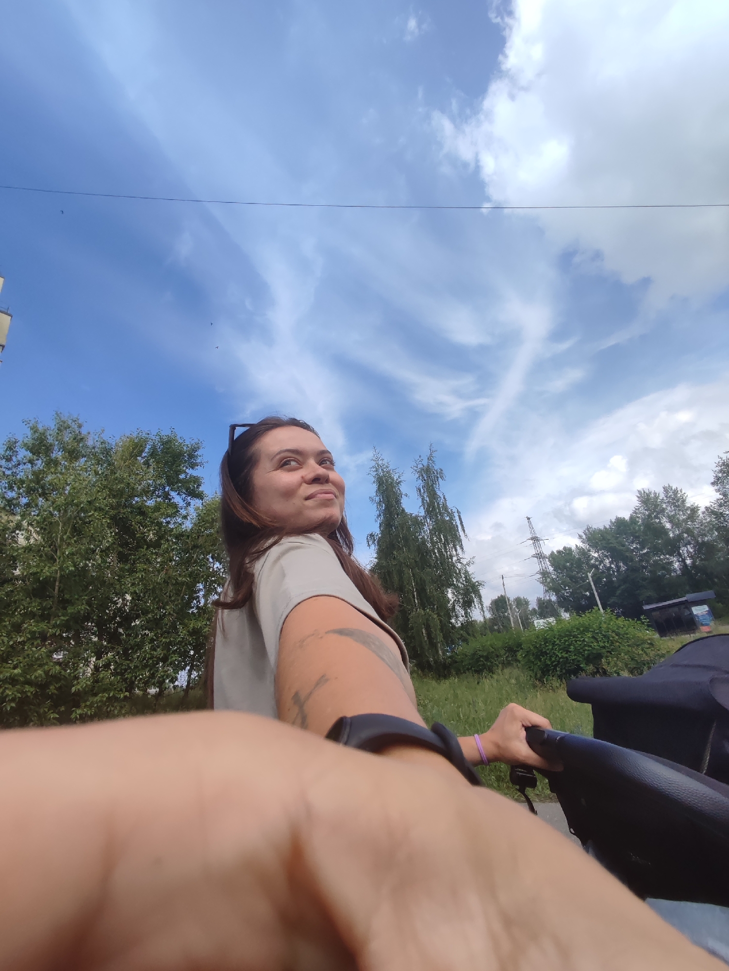 A woman is pushing a stroller outdoors in Chaikovsky, Russia. She is turned to her left, looking back over her shoulder with a slight smile. She has long, dark brown hair and is wearing a light-colored t-shirt. A tattoo is visible on her left arm. She is holding the handle of a black stroller with her right hand, and a purple bracelet is on her left wrist. In the background, there are trees, a utility pole, and a cloudy blue sky with some birds in flight. A portion of a building is visible on the far left. The scene appears to be a park or a street lined with trees.