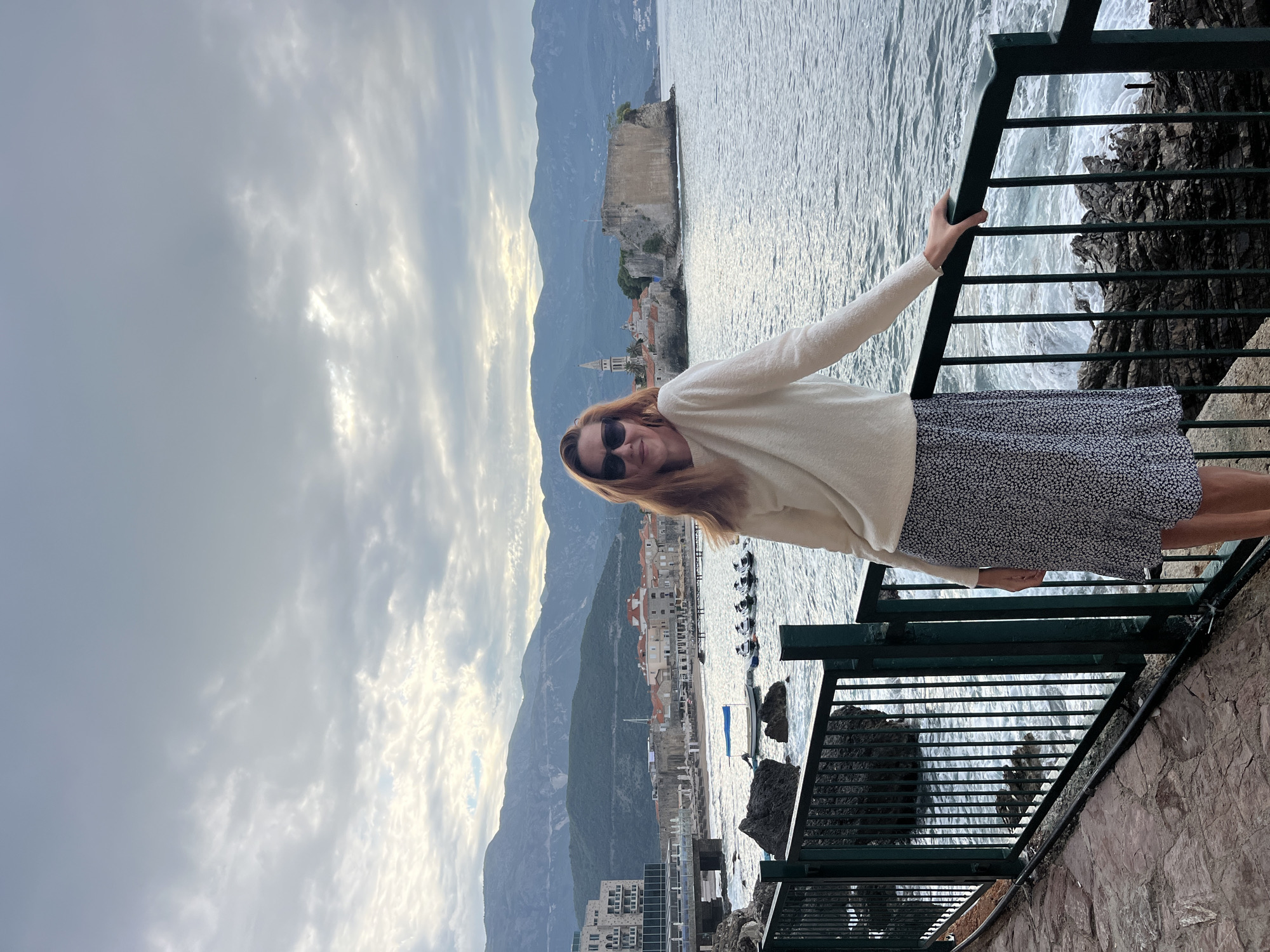 The photograph, oriented vertically, captures a woman enjoying a scenic coastal view. In the foreground, a woman with reddish-blonde hair, wearing sunglasses, a cream-colored long-sleeved top, and a dark patterned skirt, stands facing the camera. She holds onto a dark green metal railing with her right hand and displays a subtle smile, appearing relaxed. The railing separates her from a rocky cliff and the sea below, where gentle waves are visible.
In the midground, across the tranquil waters of the Adriatic, lies a picturesque coastal town, identified as Budva Old Town in Montenegro. Prominent features include a church tower with a pointed spire, ancient stone walls, and numerous traditional red-roofed buildings nestled along the shoreline. Several small boats are dotted in the water near the town. Further out, a rocky fortified islet or peninsula extends into the sea.
The background is filled with a majestic range of dark, rugged mountains, stretching across the horizon. The sky is overcast with light gray and white clouds, suggesting a soft, diffused light, possibly from late afternoon or early morning. The overall atmosphere is calm and peaceful. No visible text is present in the image.
