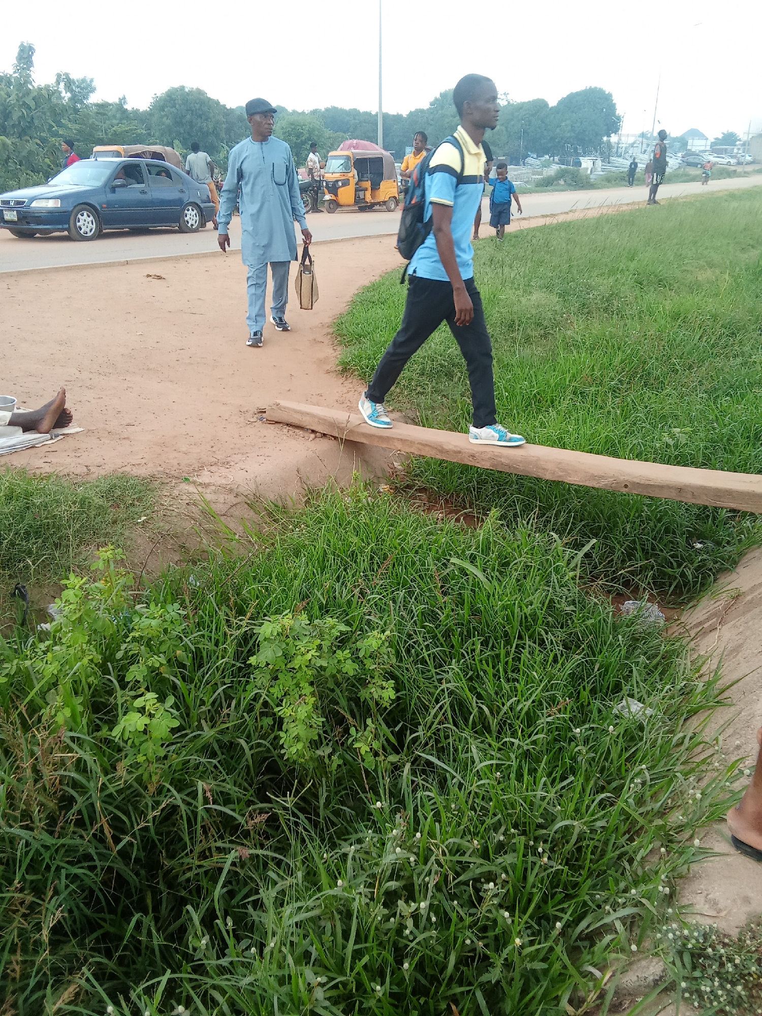 The image depicts a roadside scene in Kagini, Nigeria, under an overcast sky suggesting either early morning or a cloudy day. In the foreground, a young man wearing a blue and yellow shirt, black pants, and a backpack, is walking across a narrow wooden plank that bridges a ditch filled with lush green grass. His posture and the presence of the backpack suggest he is a student or traveler.
Further in the background, an older man in a light blue traditional attire and a cap, carrying a brown bag, walks on a dusty path adjacent to a paved road. Several vehicles, including a blue sedan and a yellow auto-rickshaw, are visible on the road, indicating it is a public thoroughfare. In the far distance, there are more vehicles and structures, hinting at a developed area. The overall setting is an outdoor environment with a mix of natural vegetation and man-made infrastructure. There is no visible text in the image.
