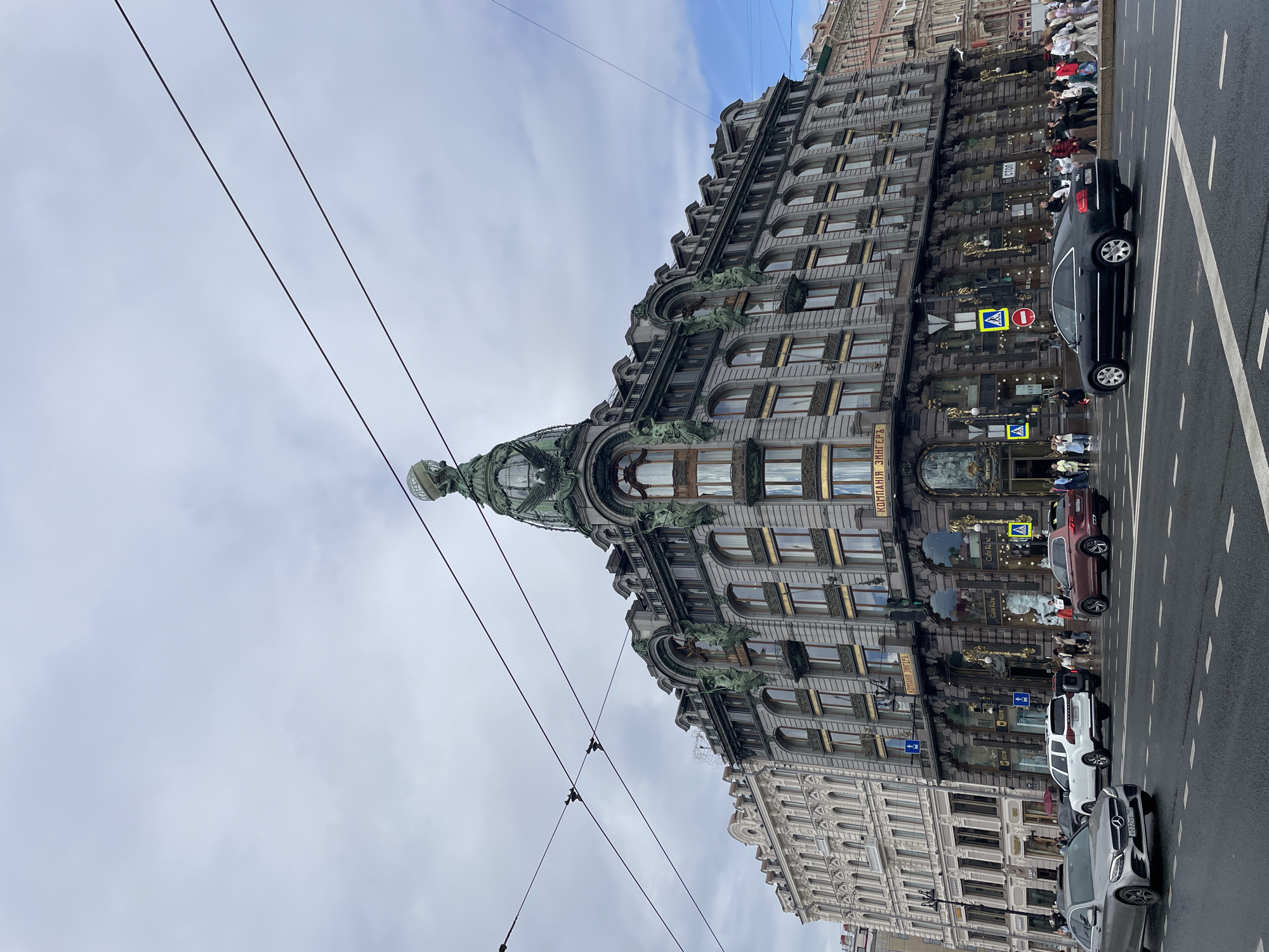 This media file depicts a street view in Saint Petersburg, Russia, focusing on the iconic Singer House (Dom Knigi). The ornate, grey stone building dominates the scene, featuring numerous windows and intricate architectural details, including sculpted figures. A distinctive green dome crowns the building's corner. The perspective is from a low angle, looking up towards the building and the sky.
Several cars are visible on the wet asphalt road, suggesting recent rain or the presence of puddles. These include a black sedan, a white SUV, and a silver car. A few people are walking on the sidewalk in front of the Singer House, some of them appearing to be tourists. The sky is overcast with light clouds, indicating a diffused daylight, possibly morning or afternoon.
Visible text includes signs on the building and street signs. One sign on the building reads "КОМП." This is likely an abbreviation related to the "Dom Knigi" which translates to "House of Books." Street signs with directional arrows and a yellow diamond sign with a red circle and a red line (likely a prohibition sign) are also present. The overall atmosphere is urban and suggests a typical day in a historic city center.