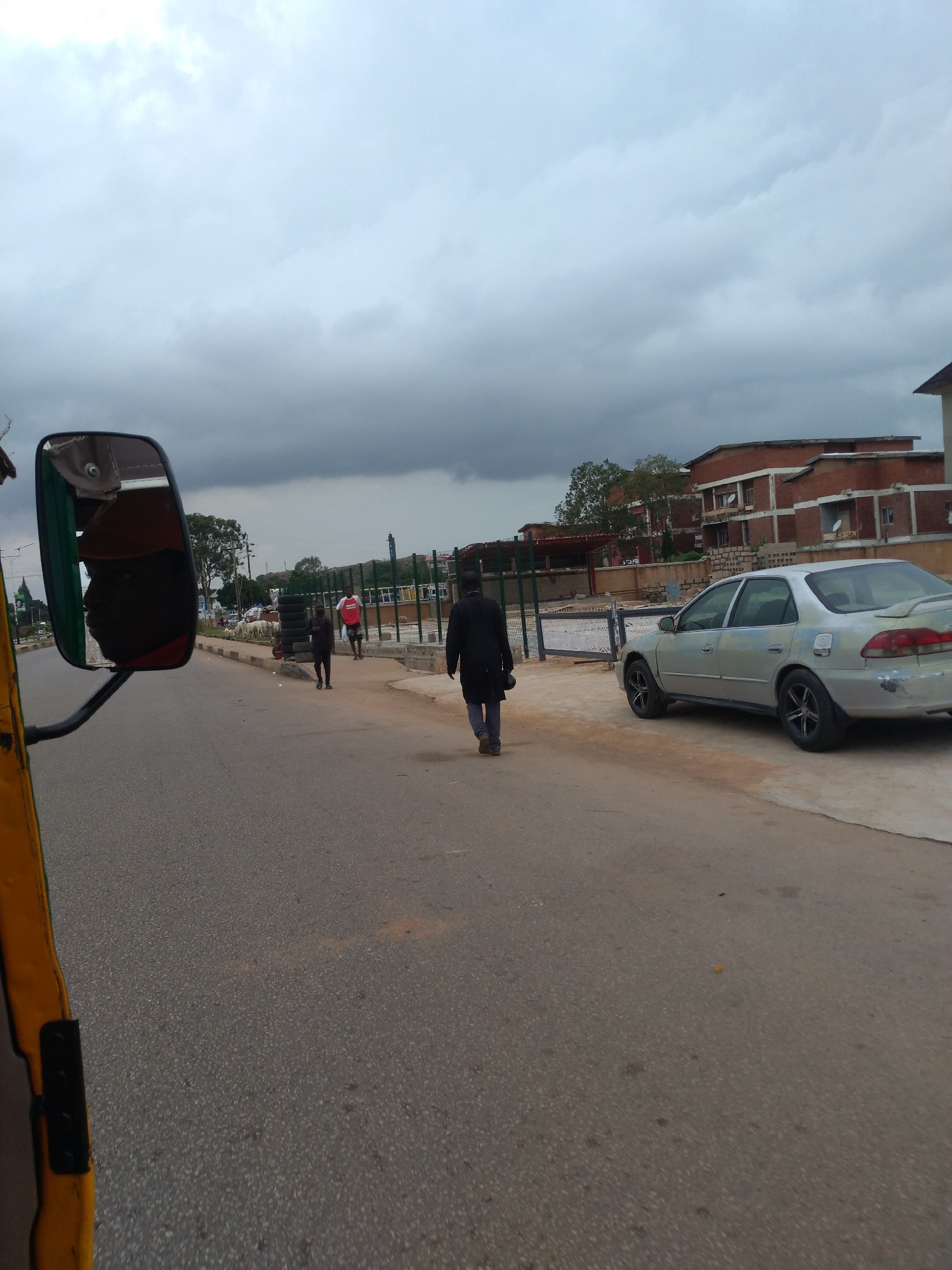 The image shows a street scene in Jos, Nigeria, on a cloudy day. In the foreground, the left side of the frame is occupied by the side mirror and part of the body of a yellow motorized tricycle, likely a 'Keke Napep'. The mirror reflects a person's face wearing a red cap and a red shirt.
On the street, there are several people walking. One man in a black coat and pants walks away from the camera towards the right side of the frame, carrying a black bag. Further down the street, a person in a white shirt and red shorts stands near a fence, and another person in a dark outfit walks away from the camera. A flock of white goats is visible on the side of the road, near a sidewalk lined with stacked tires.
In the background, there are multi-story buildings constructed with red brick and a dark gray, cloudy sky. A silver sedan is parked on the right side of the road. The road surface is asphalt.