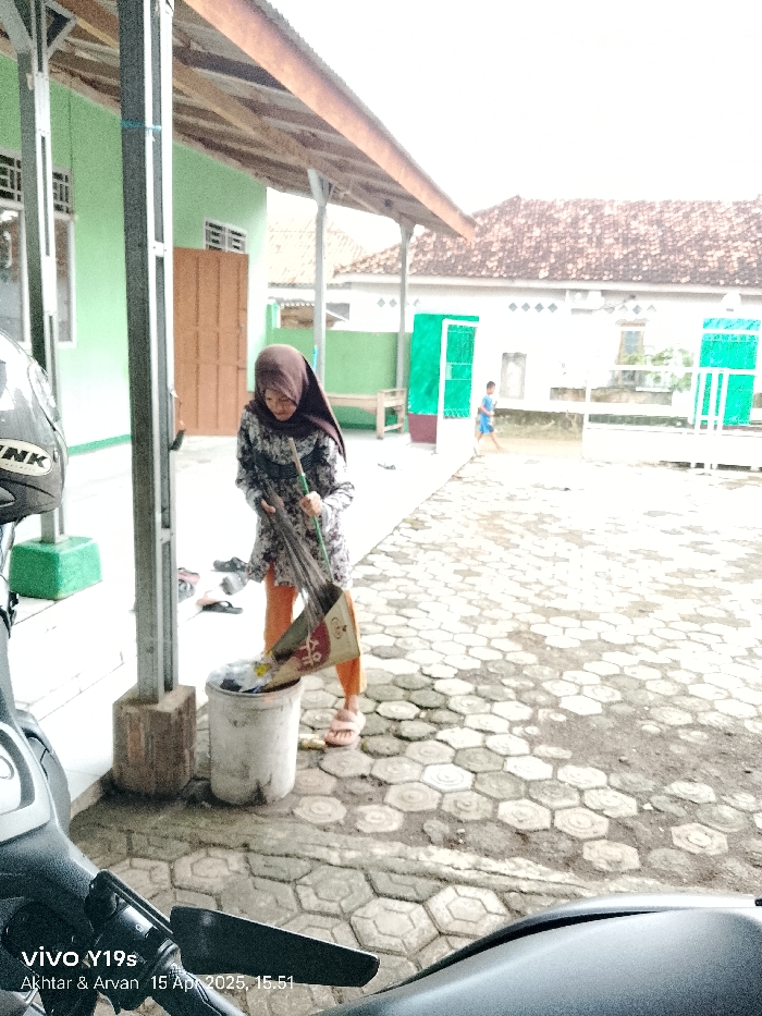 The photo, taken from the perspective of a motorcycle, shows a woman wearing a headscarf sweeping debris into a trash can. The woman is wearing an orange-and-black patterned dress. The ground is paved with hexagonal stones. There is a small boy in a blue shirt walking away from the camera in the background. The photo was taken in Anjatan, Indonesia, at the coordinates -6.419 latitude and 107.979 longitude. The image was captured on April 15, 2023 at 15:51. The woman is in the process of sweeping debris with a broom into the trash can. The photo was taken in front of a building with a green door and a tiled roof. There is a motorcycle parked in front of the building. The building is covered in a green paint.
