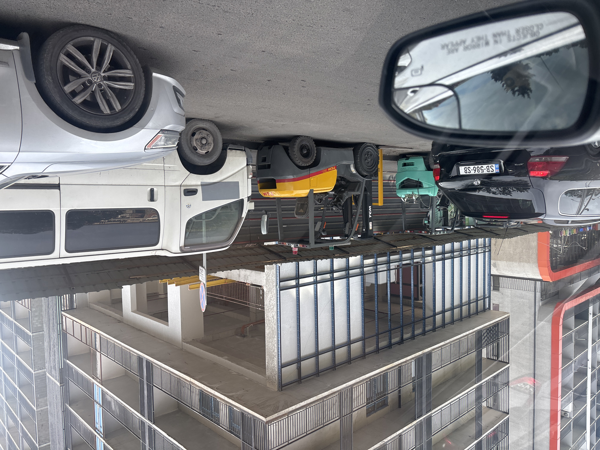The image shows a street scene in Tbilisi, Georgia. The photograph appears to be taken from inside a vehicle, evidenced by the presence of a side mirror. The view includes multiple vehicles: a silver Volkswagen, a white van, a yellow forklift, a teal utility vehicle, and a black Volkswagen Jetta with Georgian license plate SB-586-SB. In the background, there is a multi-story building under construction with exposed concrete and rebar. A “No Parking” sign is visible. The scene suggests a mixed urban environment with ongoing construction activity. The mirror indicates objects are closer than they appear.
