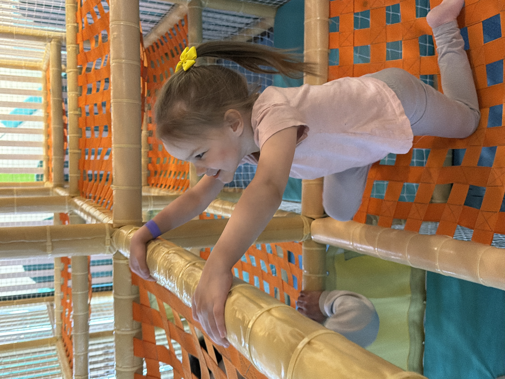A young girl wearing a pink shirt and gray pants, with a yellow bow in her ponytail, is crawling through an indoor playground structure. She is grasping a padded horizontal bar with both hands. The structure is made of beige-colored padded bars and orange netting. The background includes various levels and structures of the playground, with glimpses of blue and green padded surfaces. A part of the child's leg is visible behind the girl and in front of her. There is no specific indication of this location being in the city of Izhevsk, Russia.