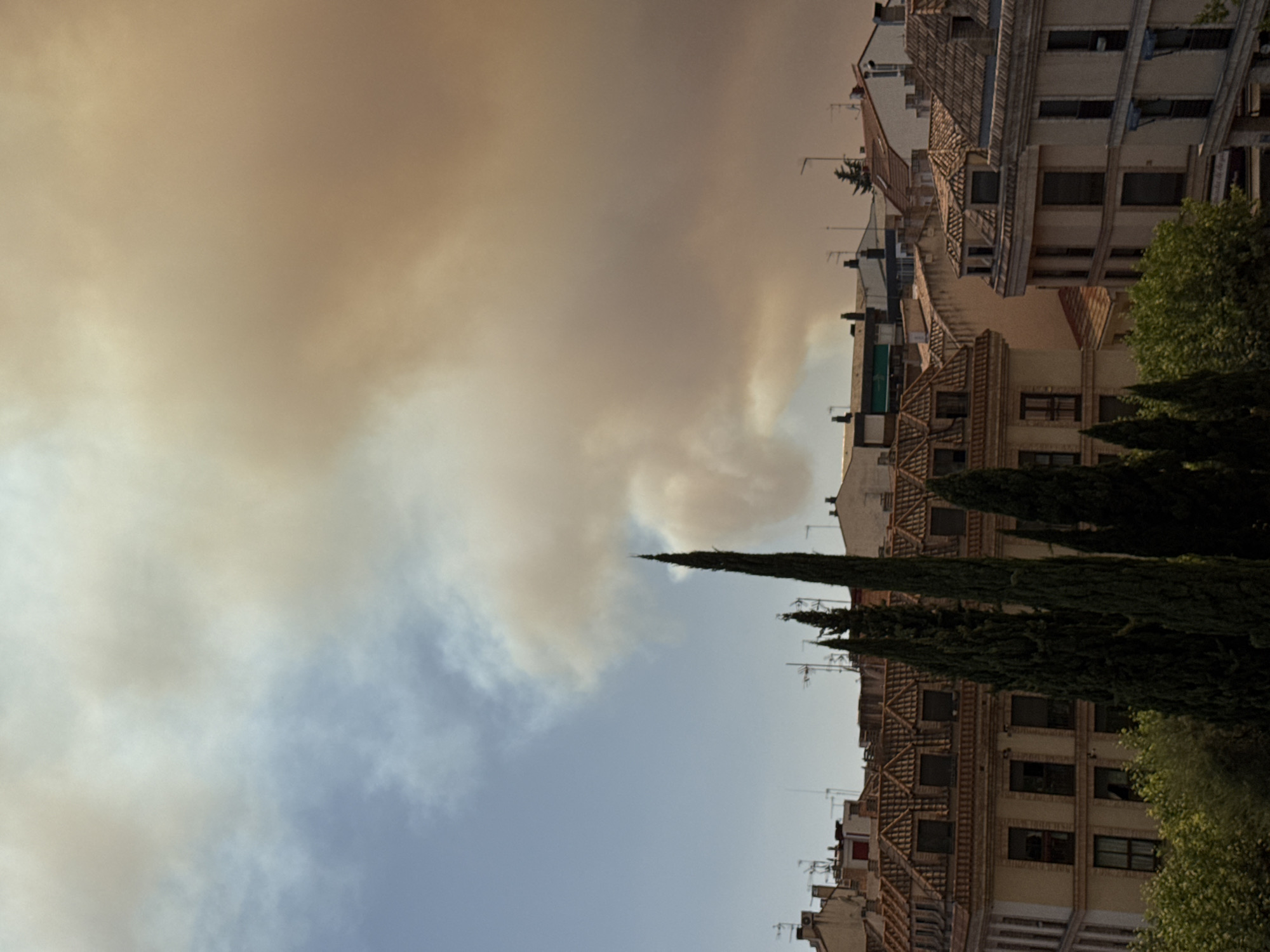 The image depicts a cityscape under a sky heavily obscured by thick, reddish-brown smoke. The smoke plumes dominate the upper portion of the image, casting an orange hue over the scene. Below the smoke, a clear blue sky is visible on the left. The middle ground is filled with the rooftops of buildings, characterized by terracotta tiles and a mix of architectural styles, suggesting a European urban environment. Some buildings have multiple stories with visible windows and balconies. Tall, dark green cypress trees are prominently positioned in the foreground, partially obscuring the buildings. The overall impression is one of an environmental event, likely a fire, impacting the atmosphere above a densely populated area. The location is specified as Las Rozas de Madrid, Spain. No people are visible in the image.