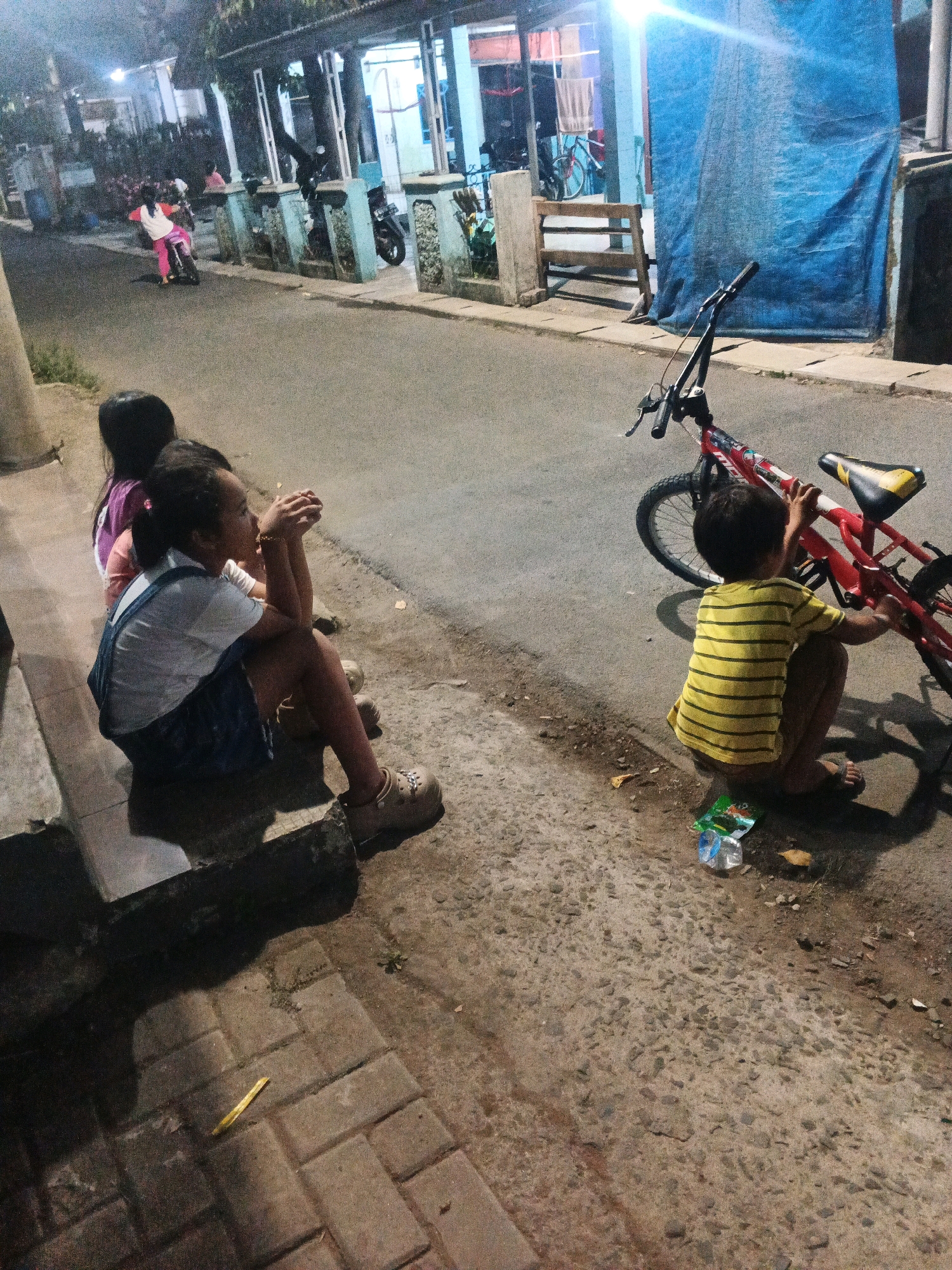 The image depicts a street scene at dusk or night in Kadu, Indonesia. In the foreground, two young girls are sitting on a low concrete step. The girl on the left has dark hair and is wearing a pink and purple shirt. The girl on the right, closer to the camera, is wearing a white shirt and blue overalls. She is looking to the right, with her hands clasped in front of her.
To the right of the girls, a young boy in a yellow and black striped shirt is crouched down next to a red bicycle. He is facing away from the camera and appears to be interacting with the bicycle. The bicycle is parked on the asphalt street.
The environment suggests a residential street. There are buildings with lights on in the background, and a blue tarp is visible covering part of a building. The street is paved with asphalt and has a sidewalk made of brick pavers in the foreground. There is some debris on the ground, including a discarded plastic bottle and wrapper near the boy and bicycle. The lighting is dim, indicating it is likely evening or nighttime. The overall impression is of children playing or resting on a street in a village or small town.