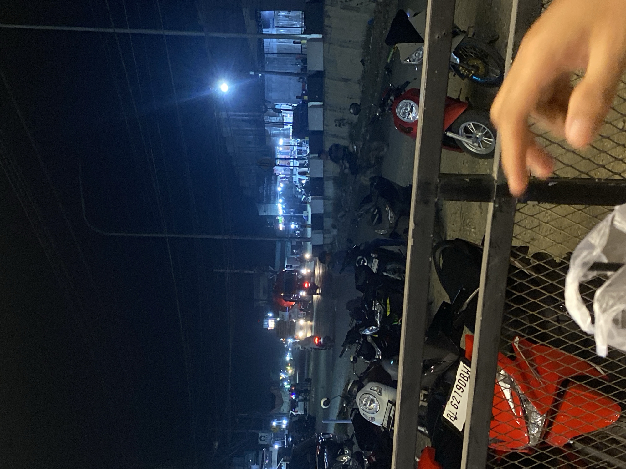 The image depicts a night scene in Tanjongkapai, Indonesia. Motorbikes are parked along a street with buildings illuminated by streetlights and storefronts. Traffic is visible in the distance, indicated by headlights and taillights. A metal fence is present in the foreground, partially obscuring the view. A hand is visible at the right of the image. A license plate BL 62190 BW is visible on one of the motorbikes.
