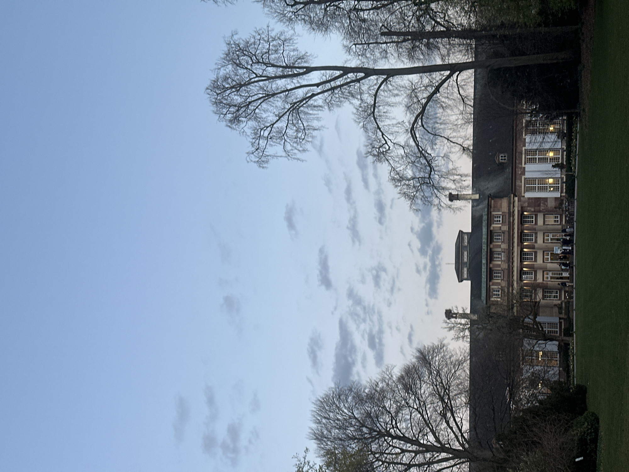 The image shows a low-angle view of a large building in Strasbourg, France, taken outdoors. The building is likely a university building, with multiple stories and many windows. The facade is made of brick and stone. In front of the building is a green lawn. Bare trees are visible on both sides of the building, framing the scene. The sky is a pale blue with scattered clouds. The image was likely taken in late winter or early spring, before the trees have leafed out. People can be seen in the distance.