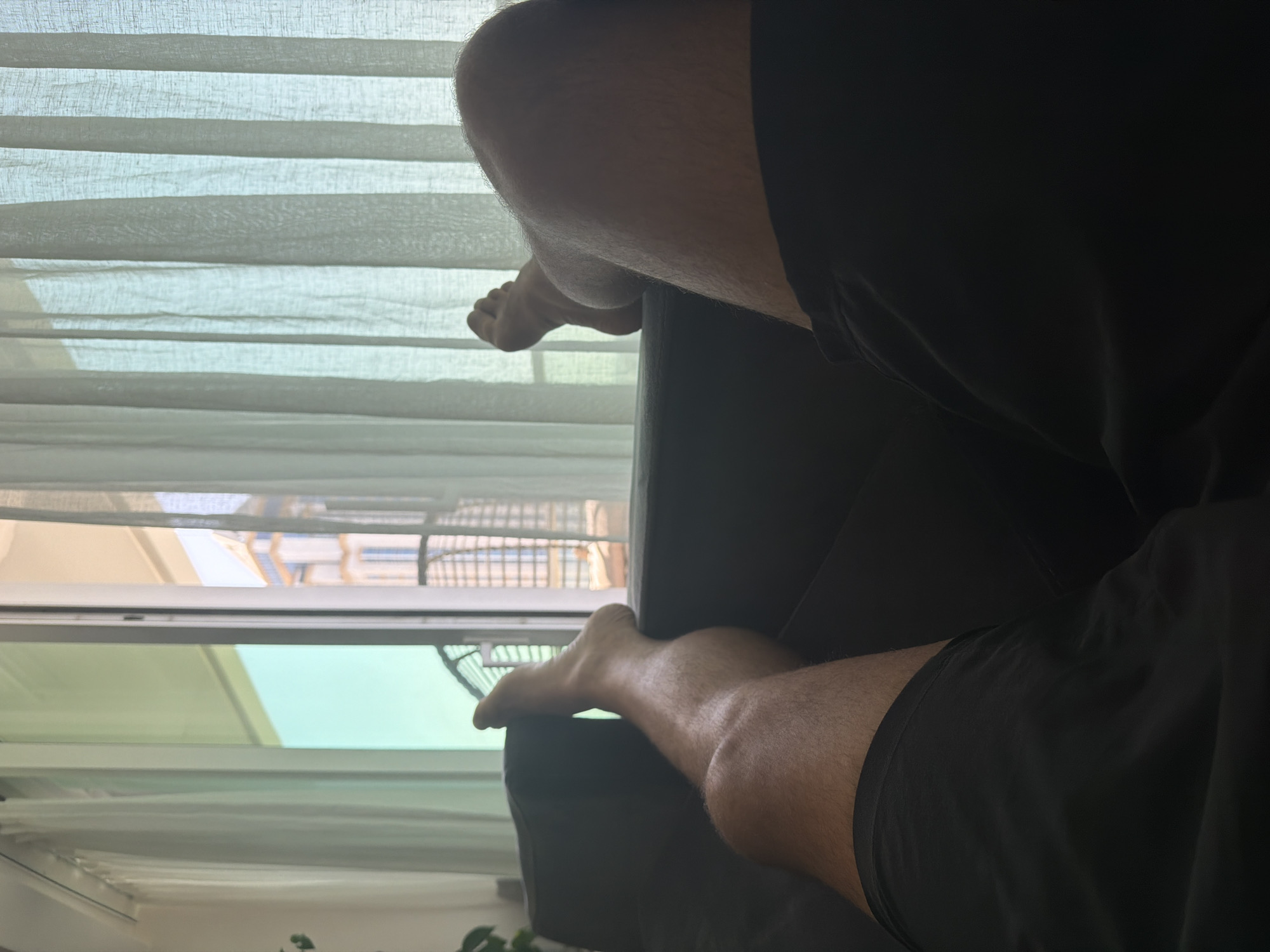 The image shows a first-person perspective of a person relaxing on a dark sofa. Their legs are extended, and they are wearing black shorts. One leg is bent at the knee, with the foot resting on the armrest of the sofa, while the other leg is extended forward. Through a glass door or window behind the sofa, sheer white curtains are visible. Beyond the curtains, a balcony railing and a building with multiple floors and windows can be seen. The overall lighting suggests it is daytime, possibly with sunlight filtering through the curtains.