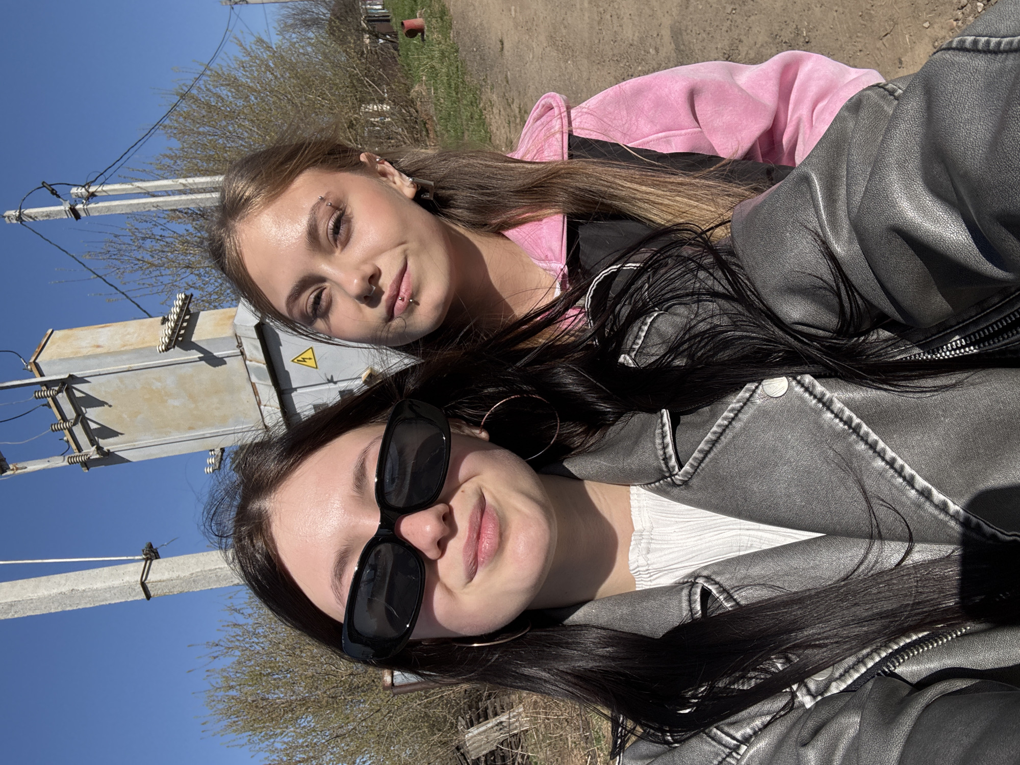 Two women are in the foreground, posing for a selfie outdoors on a sunny day. The woman in the foreground is wearing black sunglasses, large hoop earrings, a white shirt, and a grey jacket. She has long, dark hair. Behind her is a woman with light brown hair wearing a pink hooded sweatshirt and a black jacket. The woman in the back has a lip piercing. Both women are smiling slightly.
In the background, an electrical utility pole and transformer are visible on the left side of the image. There are trees with sparse foliage and a patch of brown ground. A small structure is visible in the upper right corner of the image. Given the location, the general appearance of the background aligns with typical rural or suburban areas in Фоки, Russia.