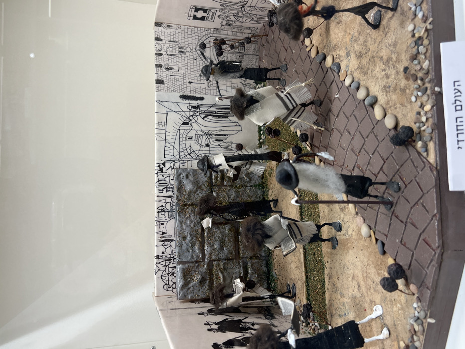 A miniature diorama, titled "העולם החרדי" (The Haredi World), is presented within a clear display case, located in the city of Yokneam Ilit, Israel. The scene depicts a gathering of stylized figures resembling Ultra-Orthodox Jewish men.
The background consists of two paper-lined walls featuring black-and-white line drawings of an ancient city, including architectural elements like arches, buildings, and city walls. A central, prominent feature of the background is a textured, three-dimensional representation of a stone wall, covered in a glossy material with gold glitter, evoking the appearance of the Western Wall.
The ground plane of the diorama is multi-textured, with a light brown, uneven, dirt-like surface occupying most of the area. A reddish-brown, tiled pathway curves through the right side of the scene, bordered by a line of small, smooth, multi-colored pebbles. Scattered across the ground are numerous small, dark, spherical, lumpy objects affixed to thin sticks, resembling stylized plants or small trees.
Approximately eleven stylized figures are visible within the diorama. These figures are crafted from dark, slender materials for their bodies and limbs, featuring textured dark brown, curly material for their heads, simulating hair or peyot. Several figures also have white, fibrous material forming beards. Most figures wear dark, wide-brimmed hats. Multiple figures are draped in white prayer shawls (tallitot) with visible black stripes and fringes. Many figures are depicted holding small, rectangular white objects resembling books, open as if being read or studied. Some figures are posed with thin, dark staffs or canes. The figures are arranged in various stances, suggesting a communal gathering, with some standing near the stone wall and others spread across the foreground. One figure on the far right appears in motion.