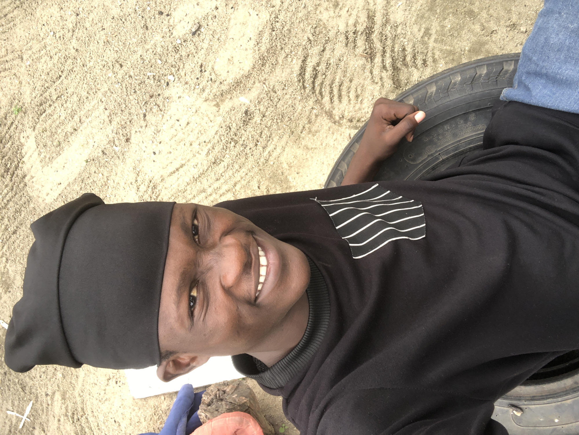 A young man with dark skin and short hair, wearing a black beanie, a black long-sleeved shirt with a white striped pocket detail, and blue jeans, is captured in a selfie. He is sitting on a tire with a weathered, sandy ground as the backdrop. The man is smiling broadly, showing his teeth, and his left hand is resting on the tire. The scene appears to be outdoors in a casual setting. The lighting suggests it is daytime. The location is Ado, Nigeria.