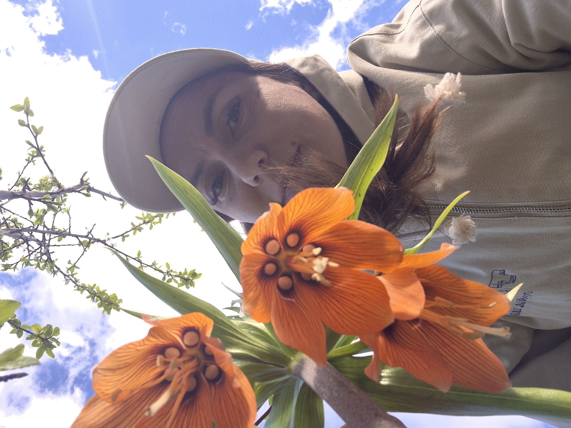 A low-angle shot captures a person wearing a beige cap and jacket. They are looking down toward the camera. Two orange Fritillaria imperialis flowers, also known as crown imperials, are in the foreground. Patches of blue sky are visible in the background. The location is identified as Chaikovsky, Russia.