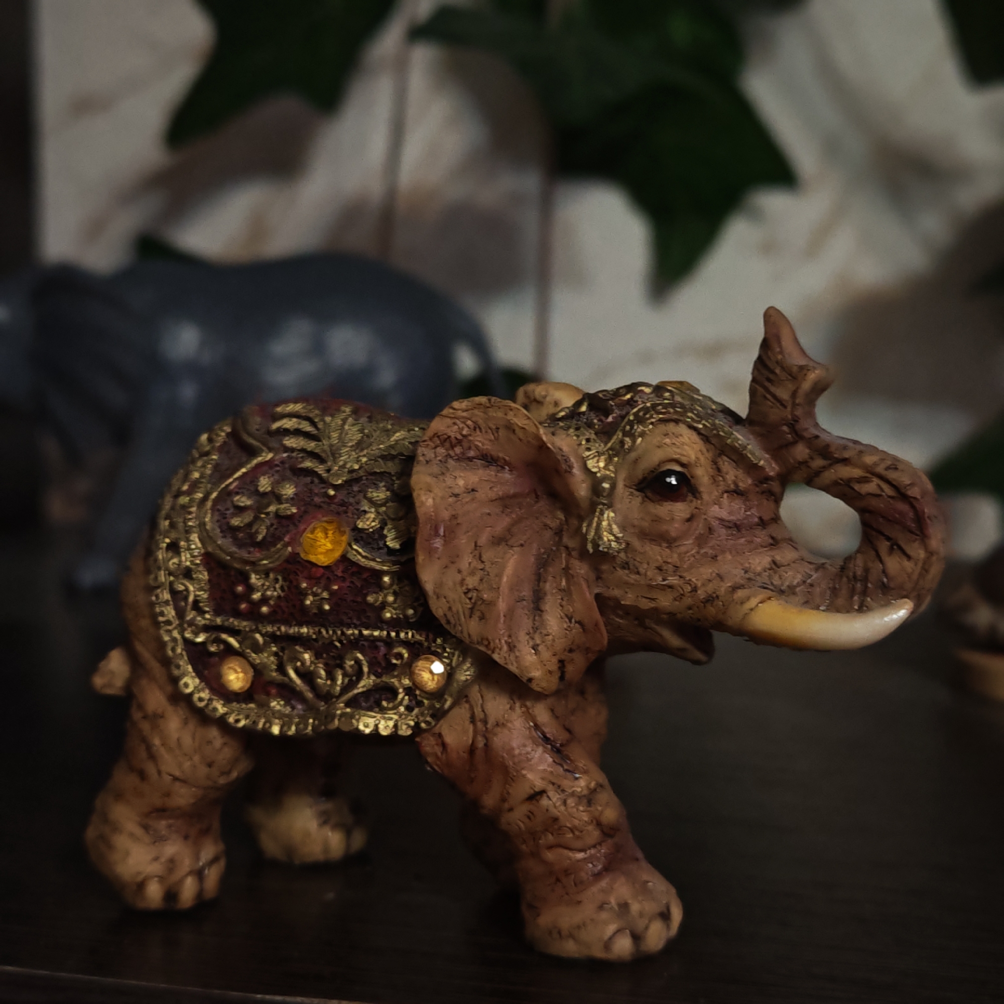 A close-up shot depicts a brown ornamental elephant figurine with a decorative blanket, positioned on a dark surface. A blurred, gray elephant figurine can be seen in the background. The background also contains blurry green vegetation and light-colored vertical lines, suggestive of wood paneling. The location in Ketoshi, Russia, is not determinable from the visual information provided.