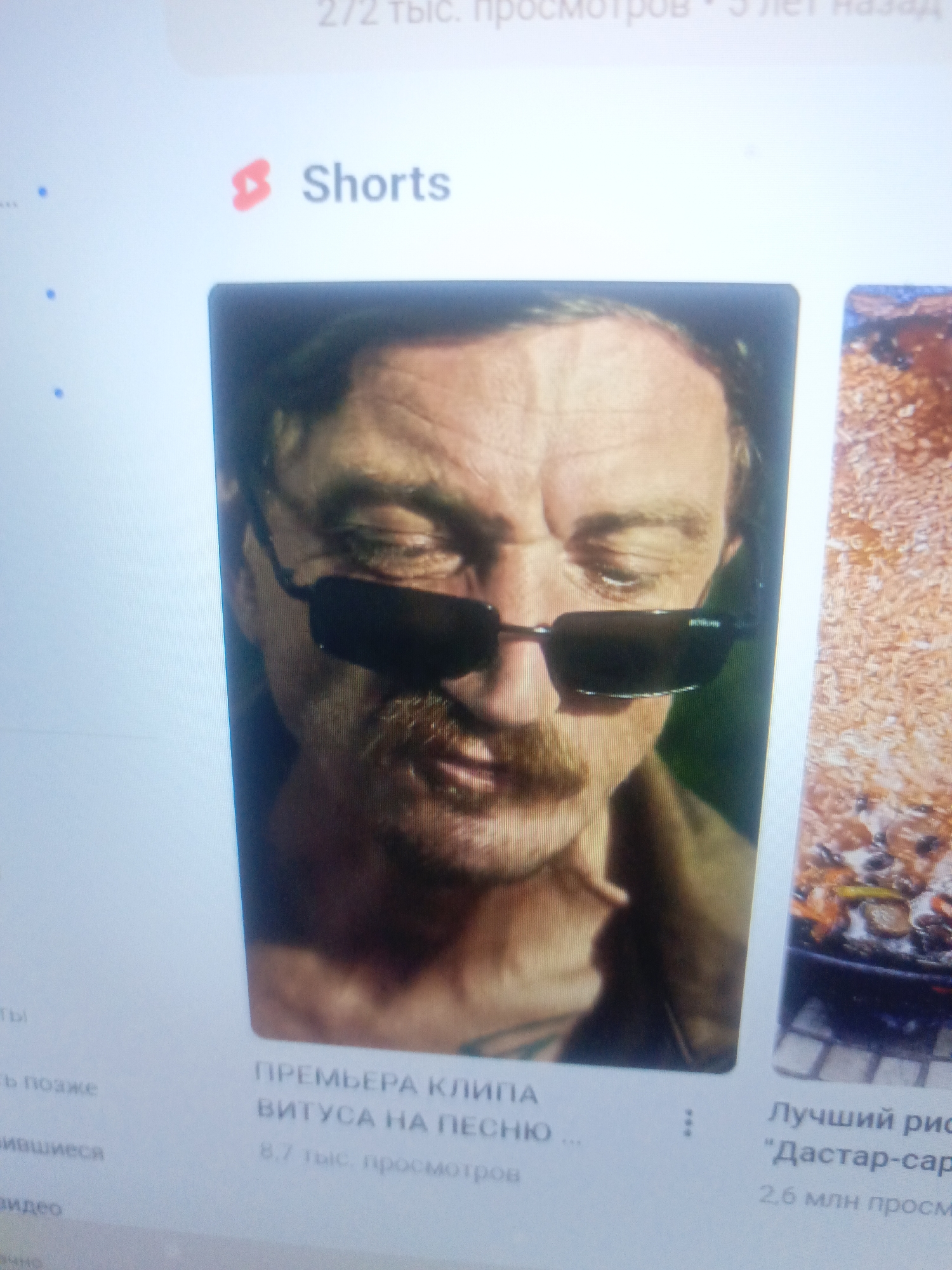 The image shows a YouTube Shorts page on a screen. The main subject is a video thumbnail featuring a close-up of a man wearing sunglasses and a mustache. Text below the thumbnail reads "ПРЕМЬЕРА КЛИПА" (Premiere of the clip) and "ВИТУСА НА ПЕСНЮ" (Vitus to the song), with "8,7 тыс. просмотров" (8.7 thousand views) below that. To the right is another video thumbnail showing food. The "Shorts" logo is visible in the top left. The image appears to be taken indoors, likely in a home or office, as suggested by the screen's reflection and the way it is captured. There is no information in the image that suggests a location in Yashkino, Russia, or any specific city.