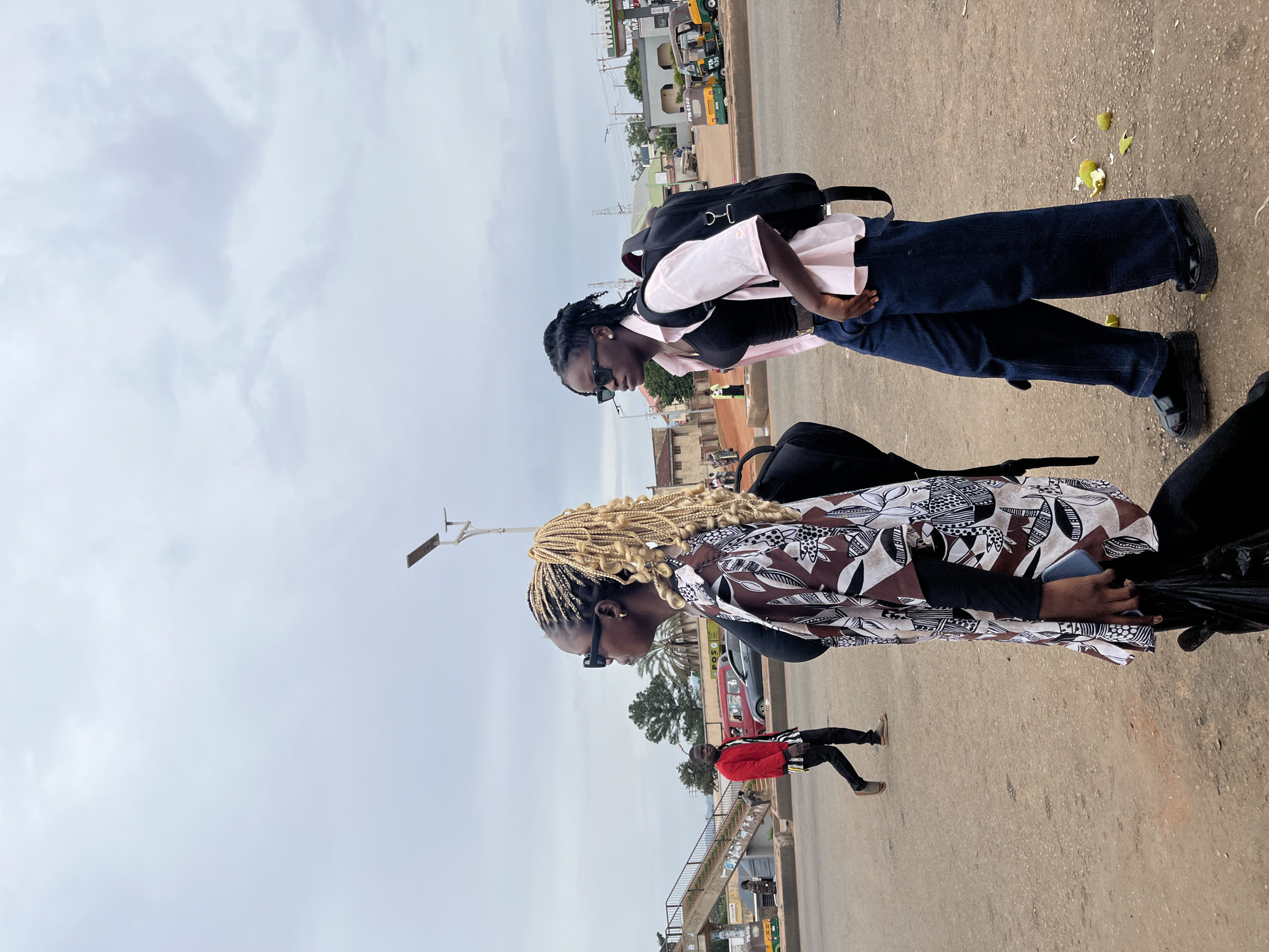 The image depicts a street scene in Gura Top, Nigeria, under an overcast sky. In the foreground, two young women are seen in profile, facing left. The woman on the left has blonde braided hair pulled into a ponytail, wears a patterned kimono-style jacket over a black top, dark sunglasses, and holds a black plastic bag and a smartphone. The woman on the right has dark hair pulled back in braids, wears a light pink button-up shirt over a black tank top, dark sunglasses, dark denim wide-leg pants, and platform sandals. She also carries a black backpack. Behind them, a man in a red jacket and striped shirt walks across the dirt road, with a pedestrian overpass visible in the background. Several vehicles, including a tuk-tuk and cars, are present in the middle ground, along with buildings and utility poles. Fallen green fruits are scattered on the ground in the lower right corner of the frame.