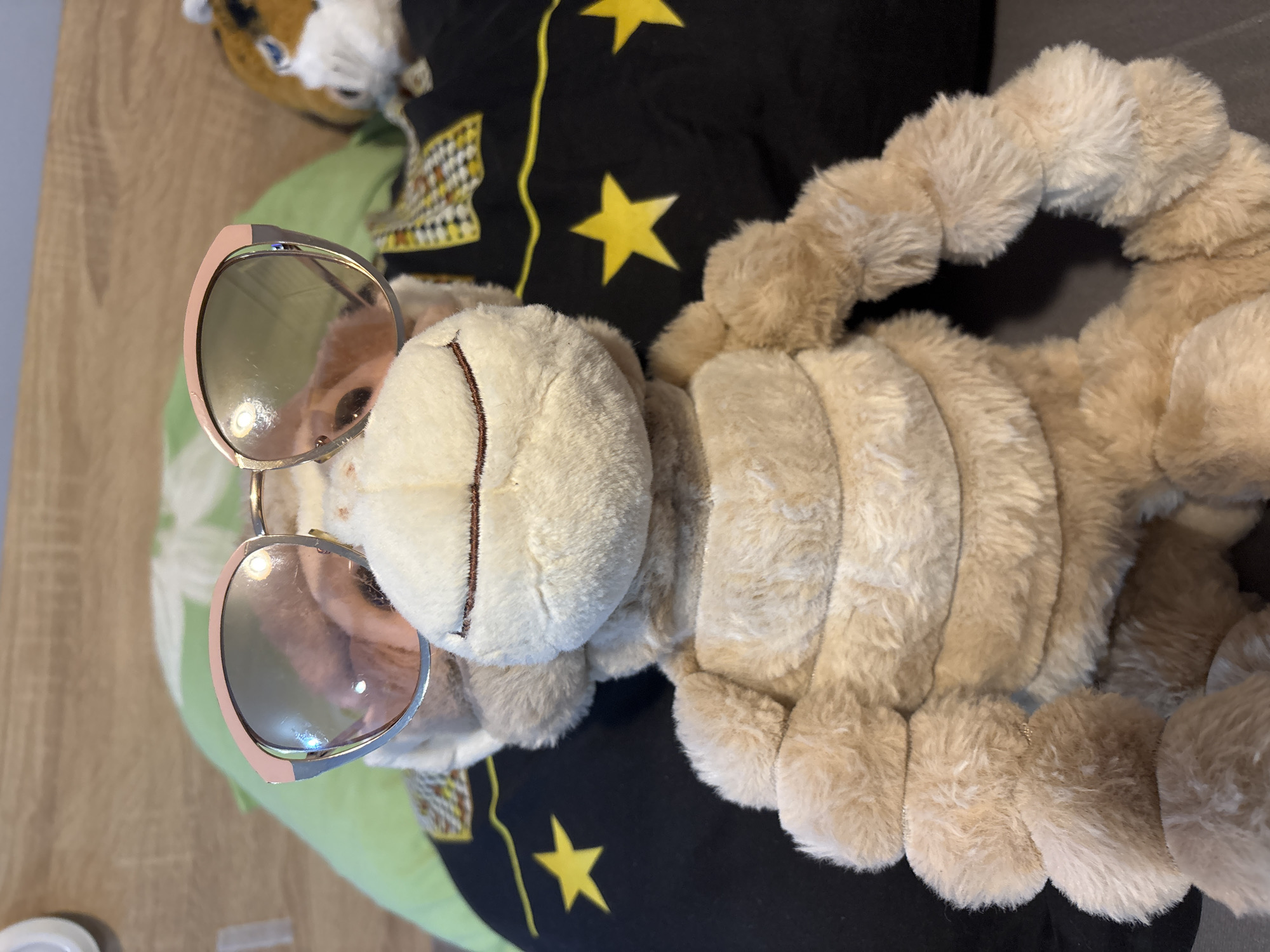 A plush, light-brown monkey toy wearing round sunglasses with pink frames is lying on a black pillow decorated with yellow stars. The monkey has a stitched smile. A green pillow is partially visible in the background, along with another plush toy. The image was likely taken indoors. The city of Sofia, Bulgaria, is not visually ascertainable from the image.