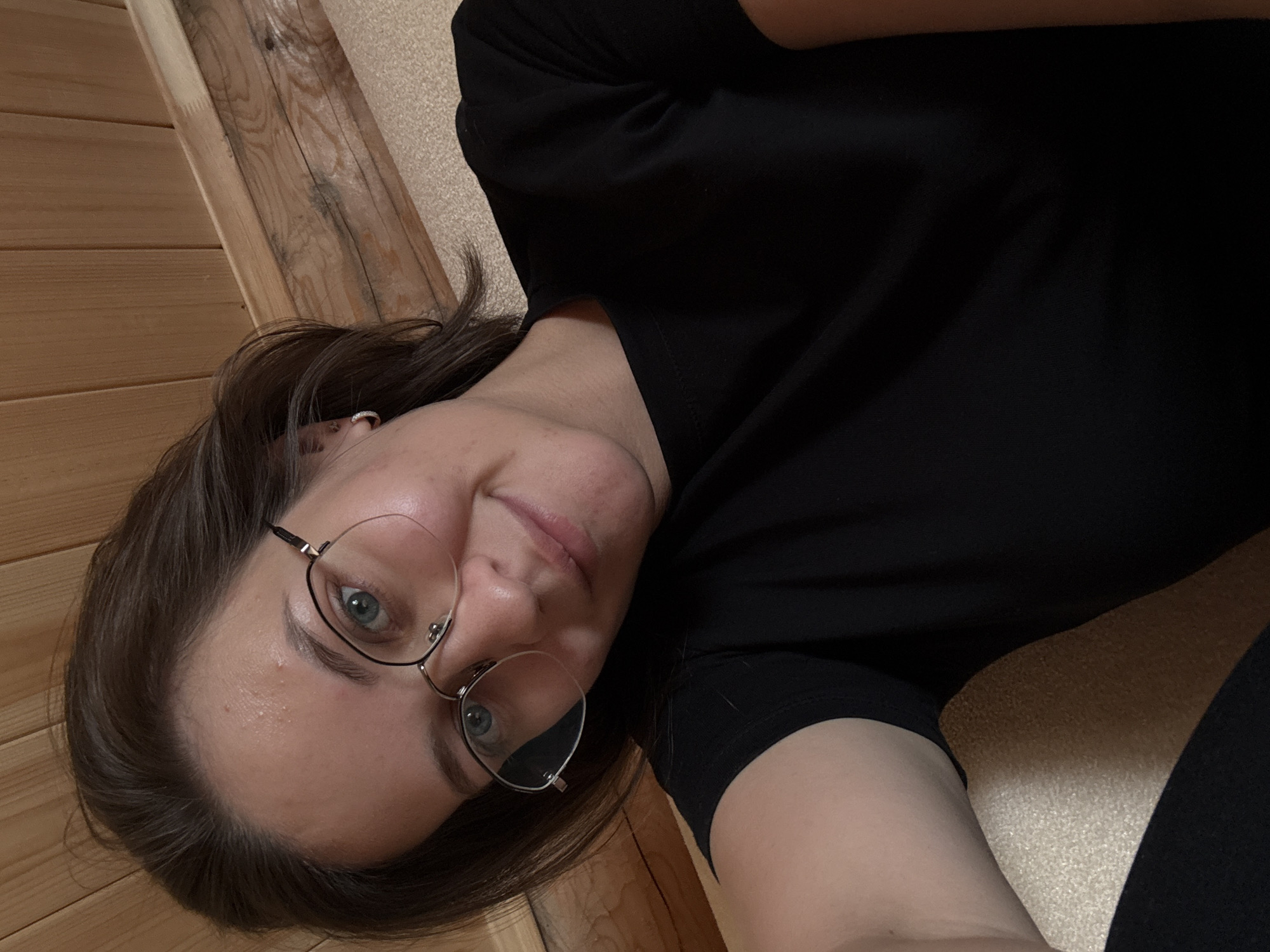 A young adult female is depicted in a self-portrait (selfie) in an indoor setting. She has medium-length brown hair, blue-green eyes, and is wearing thin-framed eyeglasses. Her expression is a subtle closed-mouth smile. She wears a plain black short-sleeved t-shirt and a small, silver-toned hoop earring in her left ear. The background features light brown wooden paneling with visible grain, suggesting a rustic or natural interior wall. A light-colored, textured surface is visible in the lower portion of the image, potentially upholstery or carpet. The image captures the subject from a slightly low angle, with her head tilted to her right. The scene is located in Lenino, Russia.