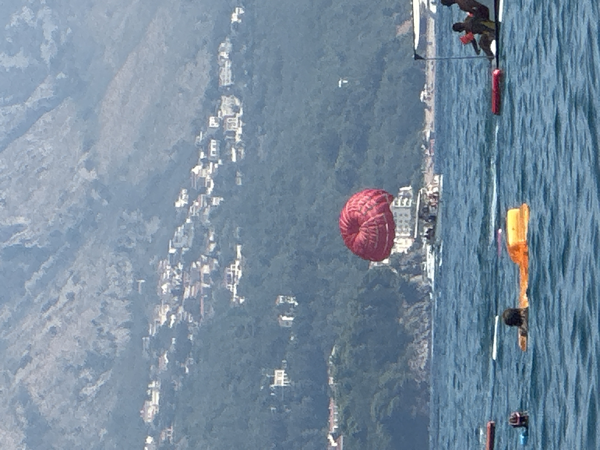 The image depicts a scene in the waters off the coast of Boreti, Montenegro. In the foreground, several people are in the water, some on inflatable floats. Two individuals are visible, one in a pink swim cap and another with dark hair, seemingly relaxing on the water. Further out, a boat is seen with two people on it, one wearing a life vest.
In the mid-ground, a boat is pulling a large red parachute, a common activity for watersports. This parachute is the focal point of the scene and is bright red. In the background, a hillside is covered in dense green foliage and dotted with numerous white buildings, indicating a town or village built into the landscape. The sea is a deep blue, with moderate waves. The sky is overcast, creating a soft, diffused light. The overall impression is a busy tourist area during daylight hours.
