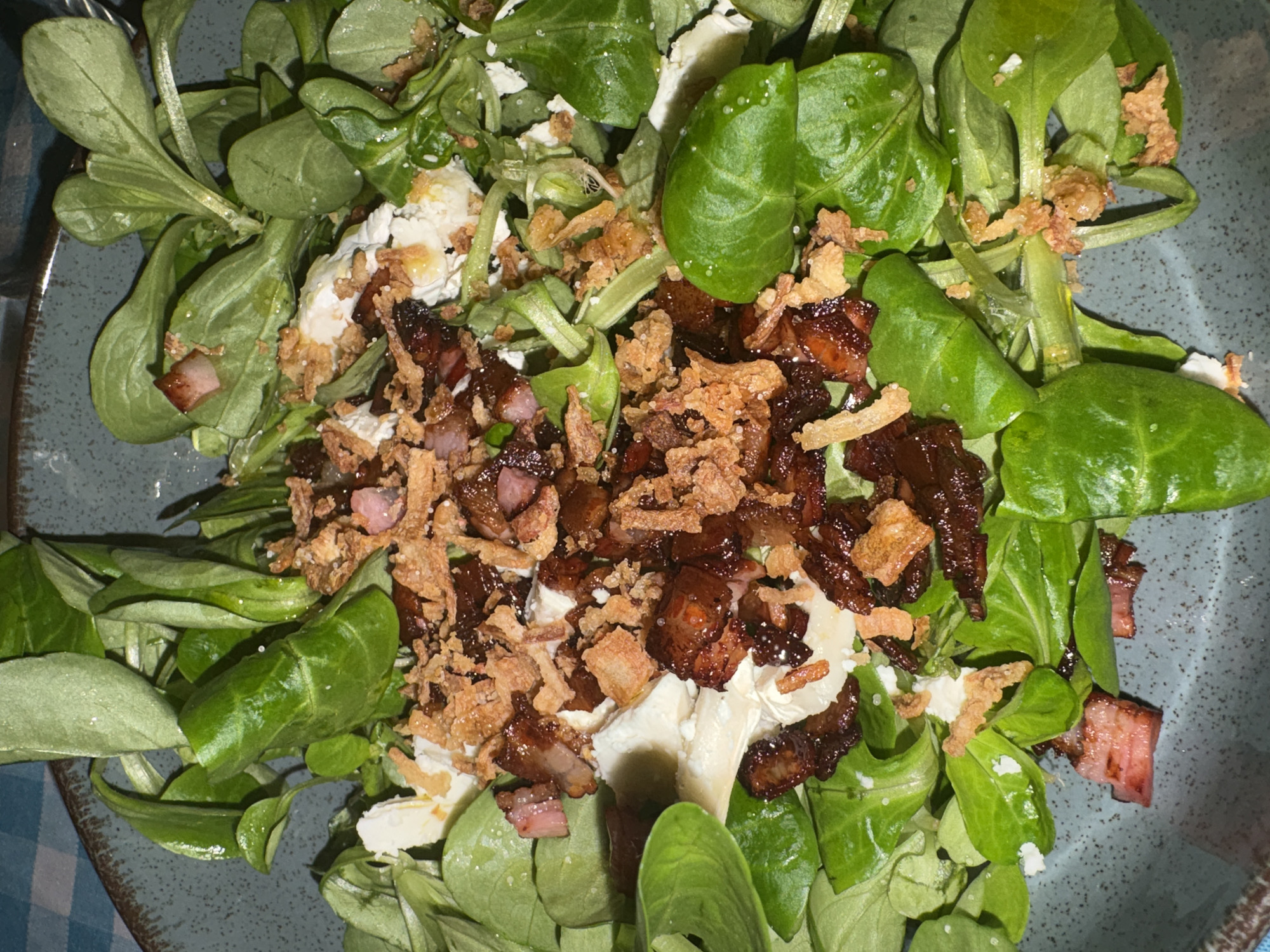 This close-up, overhead shot captures a vibrant salad served in a speckled grey-blue ceramic bowl. The main component is a generous base of fresh, bright green leafy vegetables, consistent with lamb's lettuce or corn salad (Mâche). Scattered across the greens are irregular crumbles of white cheese, possibly feta or goat cheese, and numerous pieces of cooked meat. These meat pieces vary in color from light pink to dark, caramelized brown, strongly resembling diced bacon or ham. Intermingled with the meat are abundant golden-brown, crispy bits, which appear to be fried onions or crunchy croutons, adding texture to the dish. A hint of a blue and white checkered pattern, likely from a tablecloth or napkin, is visible in the lower-left corner of the frame. The scene suggests an indoor dining setting. No people or visible text are present in the image.