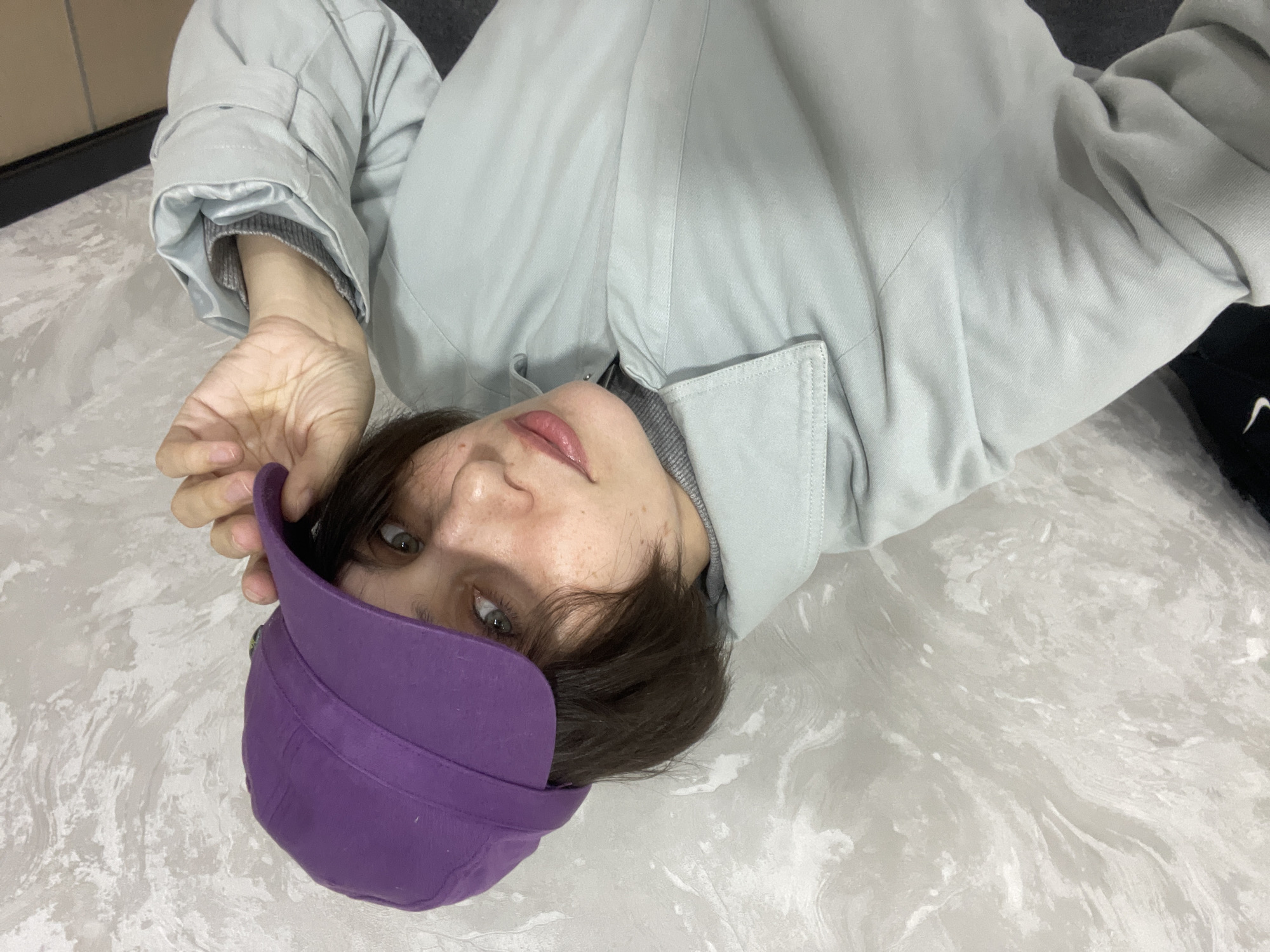 A single individual, appearing female, is depicted in a high-angle, close-up photograph. The subject has short, dark brown hair, light-colored eyes (appearing greenish-grey), and visible freckles on the face. She is wearing a grey turtleneck or sweater, layered under a light blue-grey collared jacket or coat. A purple cap, possibly a cadet or patrol style, with a flat brim, is worn on her head. Her right hand is positioned at the brim of the cap, partially obscuring the right side of her face. The subject is looking directly at the camera with a neutral expression. In the far left of the frame, a portion of a black garment with a white swoosh logo (consistent with Nike) is partially visible, likely an article of clothing or a bag. The background consists of a light-colored wall with a textured, mottled pattern in shades of grey and white. A dark horizontal line, possibly a baseboard, is visible at the bottom right, above a dark surface. The setting is indoors. No specific identifying landmarks are present to pinpoint a precise location within Izhevsk, Russia.