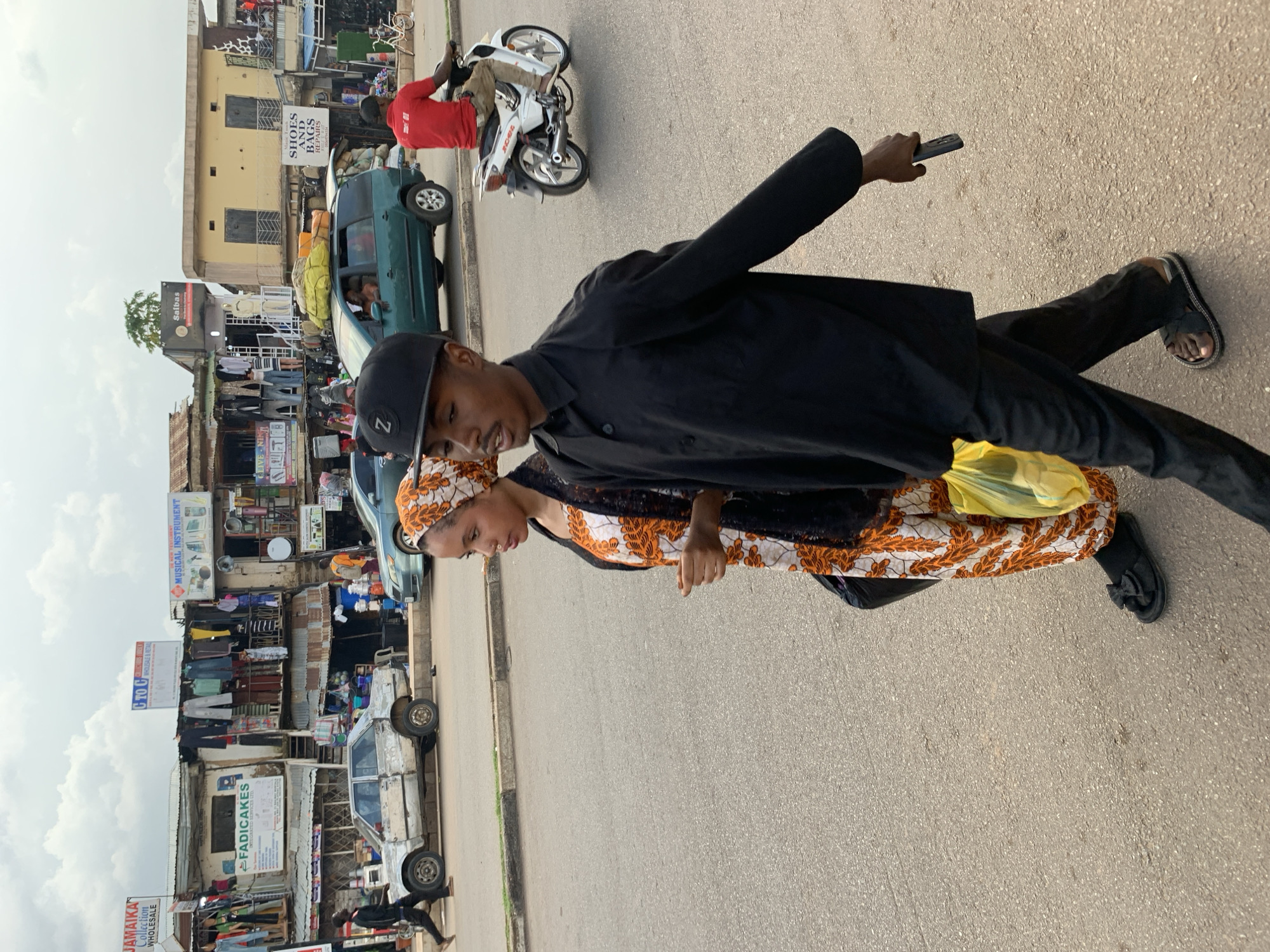 The image depicts a street scene in the city of Jos, Nigeria, likely in a commercial area with a variety of shops lining the street. In the foreground, a young man wearing a black cap and a black long-sleeved shirt and trousers walks alongside a young woman. The woman is wearing a patterned headscarf and a white and orange patterned dress with a black overlay. She is carrying a yellow plastic bag.
Further down the street, a man in a red shirt is riding a white motorcycle. Other vehicles, including a light green SUV and a white car, are visible on the road. The buildings in the background are shops with various signs, some displaying clothing and other goods. The overall environment suggests a bustling urban marketplace with people and traffic. The sky is overcast with clouds.