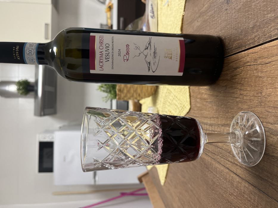 The image displays a still life captured within a residential kitchen in Ercolano, Italy. On a wooden table, a stemmed glass, exhibiting a diamond-cut pattern on its bowl and base, contains dark red wine, approximately one-third full. To the right of the glass, a dark-colored wine bottle stands, appearing mostly full. Its label reads "CANTINE ASTRONI LACRYMA CHRISTI VESUVIO Rosso 2024," and includes an illustration of Mount Vesuvius, identifying its regional origin near Ercolano. A yellow, textured placemat is partially visible beneath the bottle and extends toward the glass. The background details a kitchen area, featuring a white refrigerator with a white microwave oven positioned on top, and a stainless steel range hood above a countertop that holds a small potted plant. The scene is illuminated by interior artificial lighting. No individuals are present.