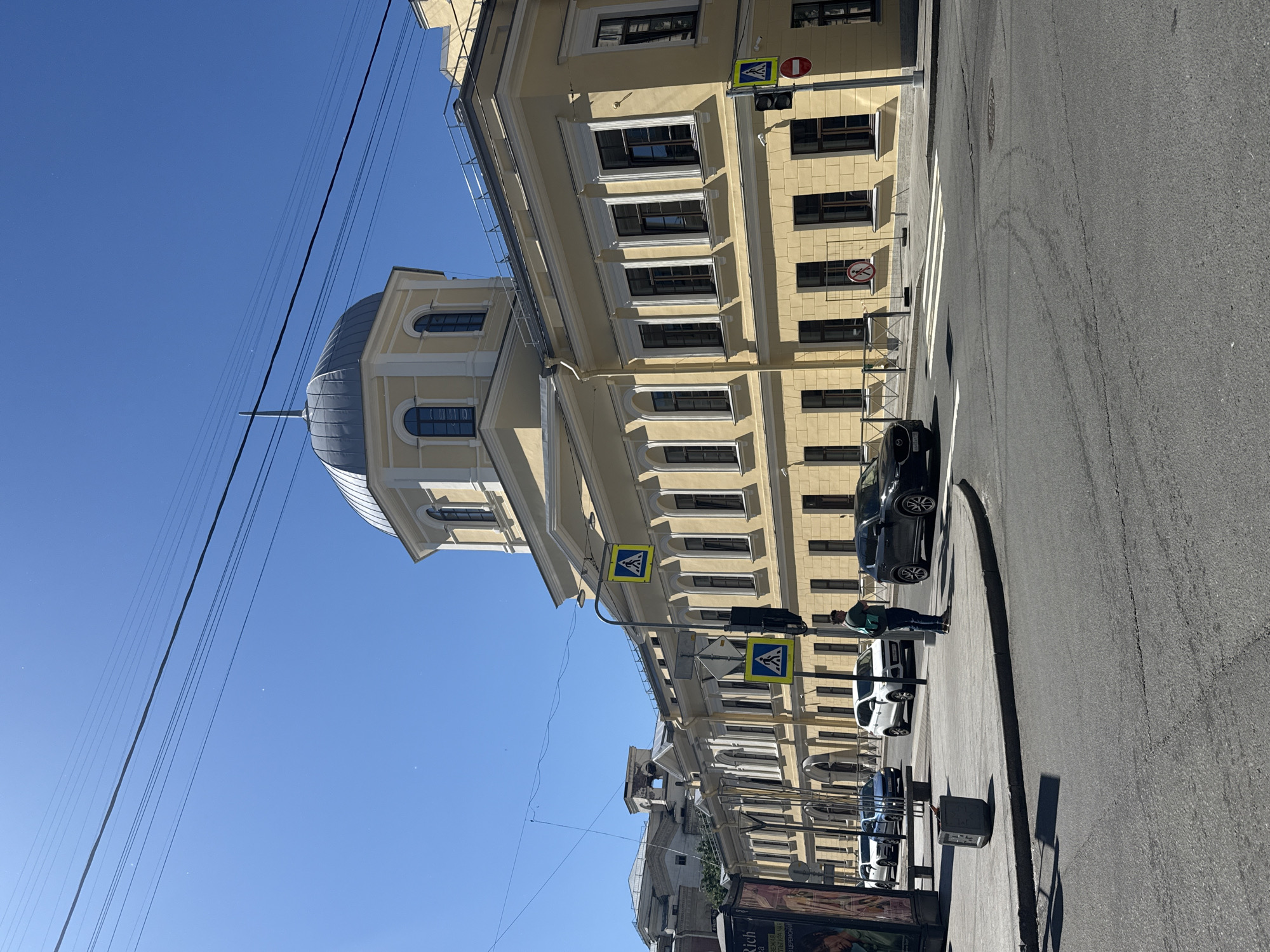 The image depicts a street scene in Saint Petersburg, Russia, under a clear blue sky, suggesting it is daytime with pleasant weather. The focus is on a light yellow, classical-style building with multiple windows and architectural details. A prominent dome tops a section of the building.
Several pedestrian crossing signs are visible on street poles, along with traffic lights. Cars are parked along the street, and a few people are present. One person, wearing a green jacket and blue jeans, is standing near the sidewalk. Another person is visible in the distance. The street itself is asphalt, with visible tire marks and painted white lines indicating a pedestrian crossing.
The scene appears to be a typical urban environment. The architecture of the building suggests a historical or cultural landmark. A partially visible advertisement is in the bottom right corner, but the text is not legible enough to decipher. The overall impression is one of a bright, sunny day in a city with well-maintained infrastructure.