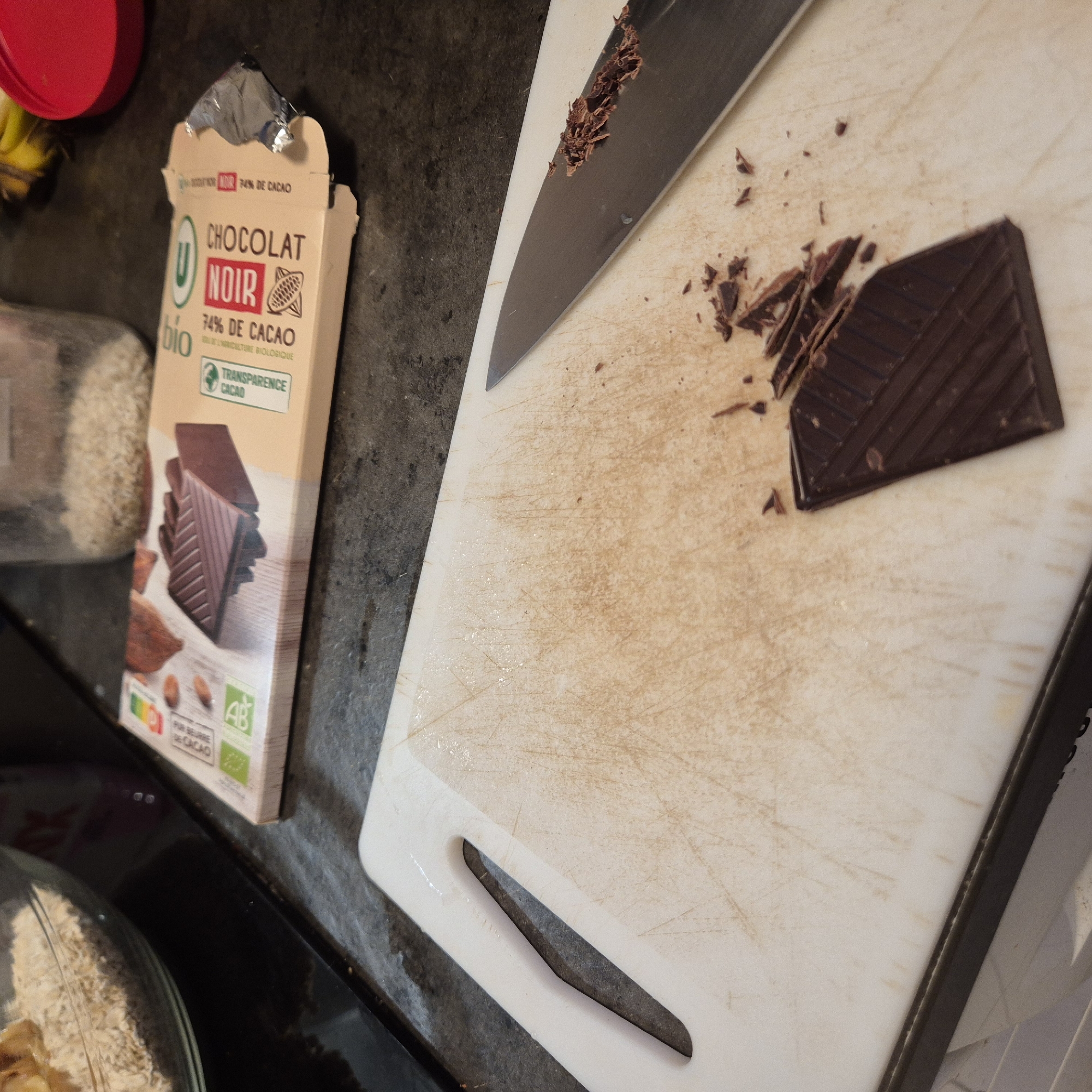 The image shows a box of U bio Chocolat Noir 74% cacao next to a white cutting board with a knife and partially chopped chocolate. The scene is indoors, likely in a kitchen. Other visible items include a red jar and a bowl with food. The scene provides no information about the location being Strasbourg, France.