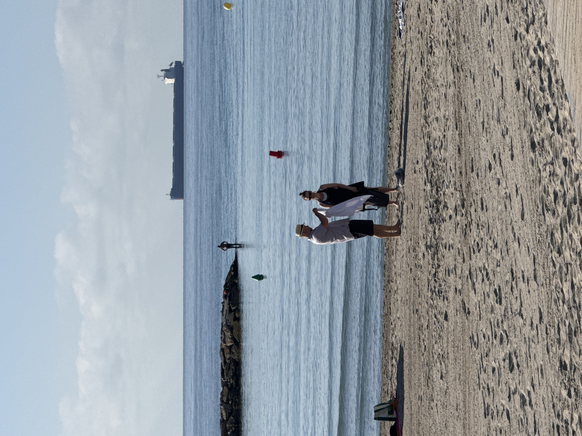 Two people, a man and a woman, stand on a sandy beach. The man, wearing a light-colored short-sleeved shirt, dark shorts, and a straw hat, holds a white cloth. The woman, wearing a black dress and sunglasses, stands next to him and also holds a white cloth. They appear to be interacting with the cloths. In the background, the calm blue ocean stretches to the horizon, dotted with a few buoys and a large cargo ship. A breakwater made of large rocks is visible in the middle ground, with a person standing in the water near it. The sky is clear with a few scattered clouds. The beach itself shows footprints in the sand, and a bag is placed on the sand to the left.