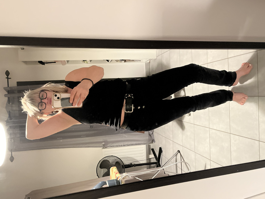 A woman in black jeans, a black top, and glasses stands in a room, taking a mirror selfie. Her blonde hair is short and messy. She wears a black bracelet on her left wrist. A black belt with silver accents is visible around her waist. In the background, there's a white radiator, grey curtains, and a black fan. The room has light-colored tile flooring. The mirror has a thick black frame.