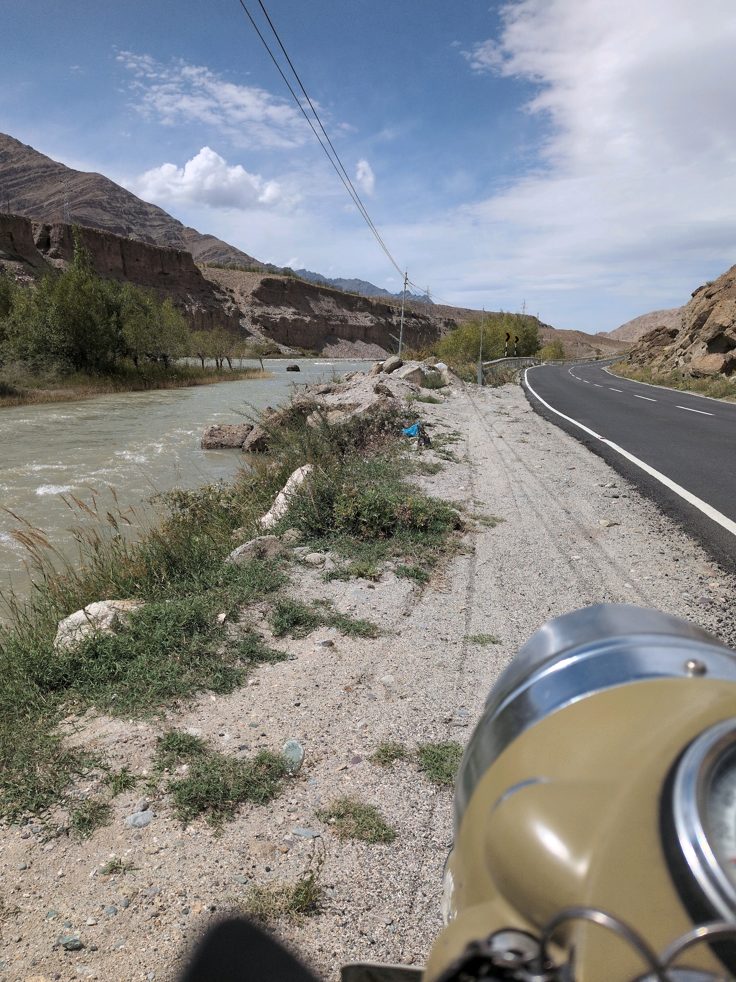 A motorcycle's tan-colored headlight and part of its body are in the foreground, facing right. The motorcycle is on a gravelly shoulder next to a paved road. The road curves to the right, disappearing into the distance. Mountains with layered rock formations line the left side of the road, where a wide, murky river flows. Sparse green vegetation grows along the riverbank and on the slopes of the mountains. The sky is blue with scattered white clouds. There is no visible sign of people or any interaction. The scene appears to be in a natural, arid environment, likely a roadside in Upshi, India, given the typical landscape.