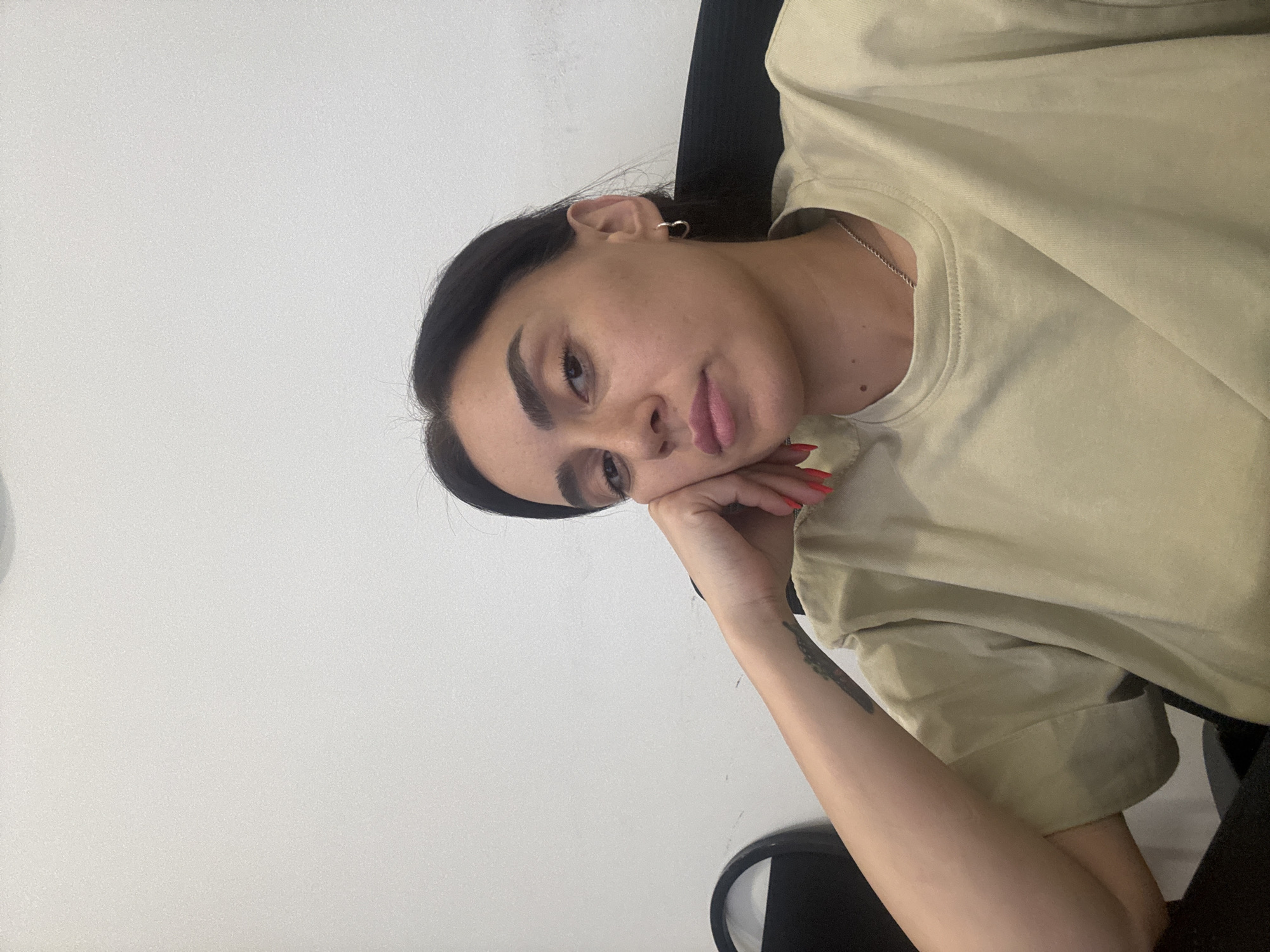 A young woman with dark hair and well-defined eyebrows leans her head on her hand, resting her cheek against her palm. She is wearing a light beige t-shirt and a silver necklace. Her fingernails are painted a bright red. A subtle tattoo is visible on her forearm. The background is a plain, light-colored wall. The image has a casual, indoor feel, with soft lighting. The woman's expression appears pensive or relaxed.