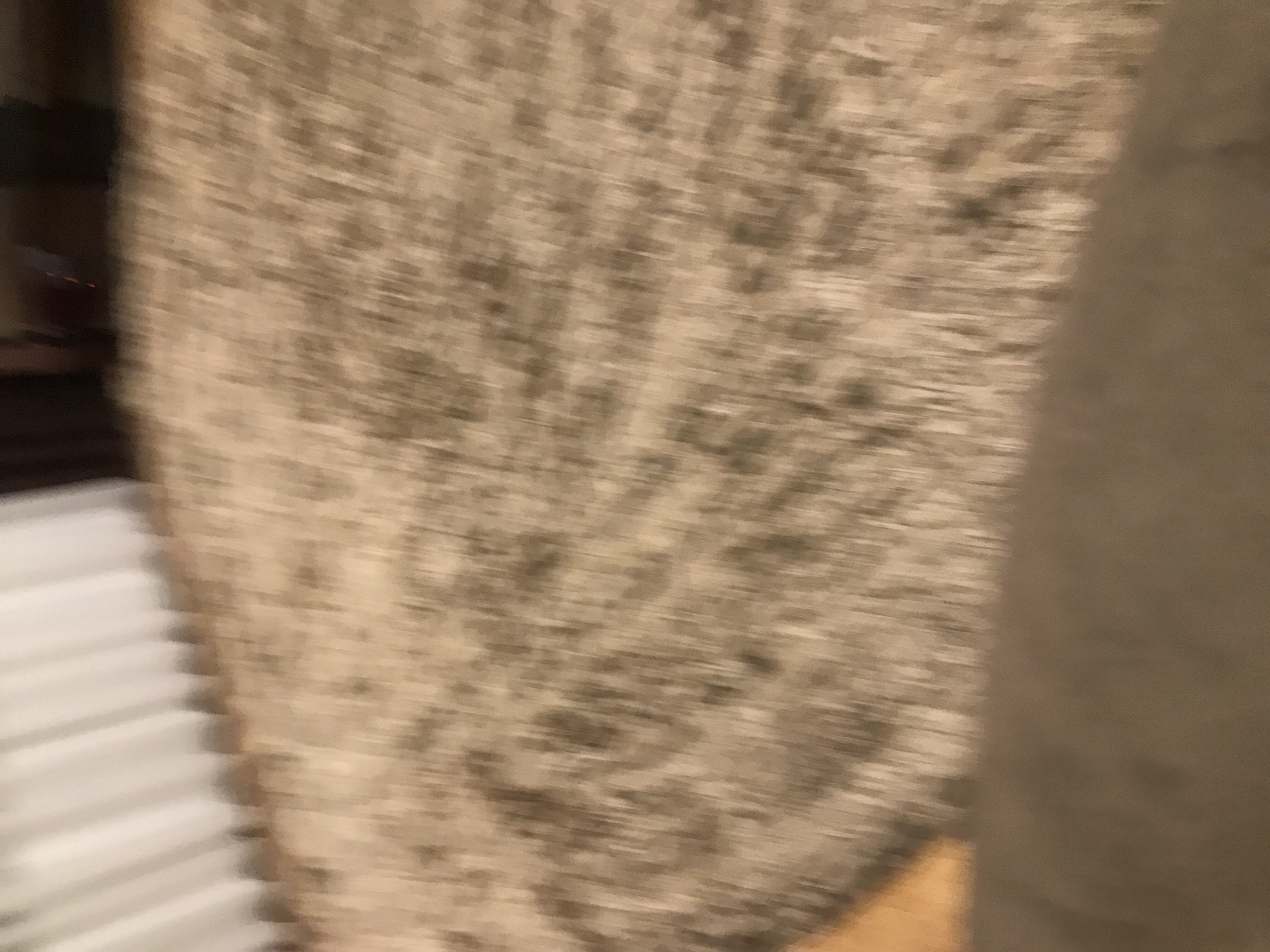 The image is blurry and out of focus, but it appears to show a portion of a room. On the left side, there are white, ribbed structures that could be blinds or part of a radiator. The central area is filled with a patterned, beige and brown texture that is most likely a rug or carpet. On the right side, there is a large, solid brown shape, possibly a wall or a large piece of furniture. The overall lighting seems dim. The location is not discernable from the image and therefore cannot be identified as Yerevan, Armenia.