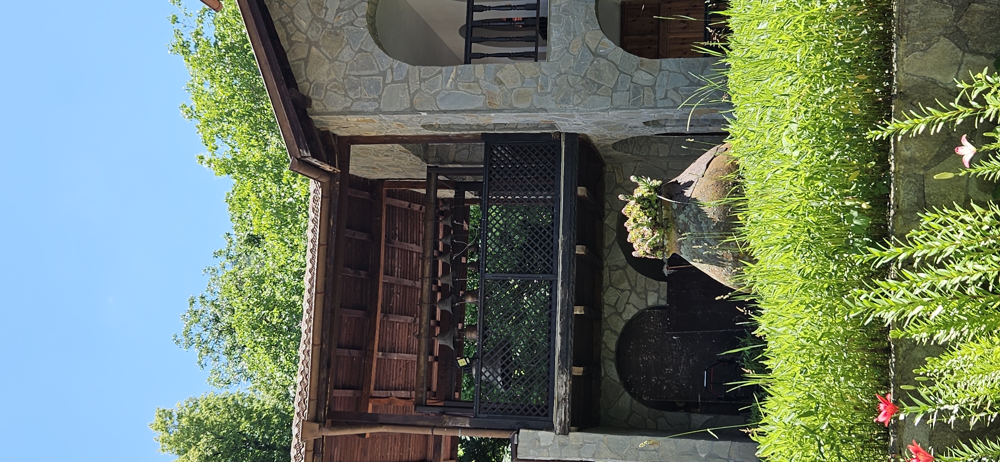 The image captures a sunny day scene at a building with stone walls and a wooden-covered balcony featuring a row of bells. In the foreground, lush green grass and some red lilies are visible. Behind the grass, a large, weathered ceramic pot serves as a planter for green foliage. The building has arched openings and wooden doors on the lower level. The overall environment appears to be outdoors, with trees and a clear blue sky in the background. The scene is located in Ovchartsi, Bulgaria.