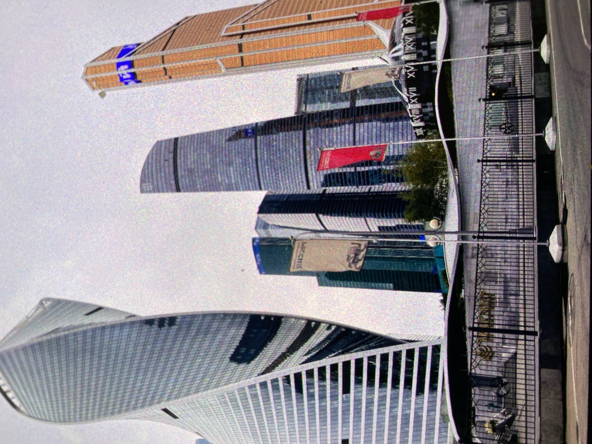 The image shows a view of the Moscow International Business Center in Moscow, Russia. The image is rotated 90 degrees clockwise. The scene includes several skyscrapers with distinct architectural styles. A metal fence runs along the edge of a walkway and road. Flags with inscriptions are visible on some of the buildings. The sky is overcast, providing a muted light to the scene.