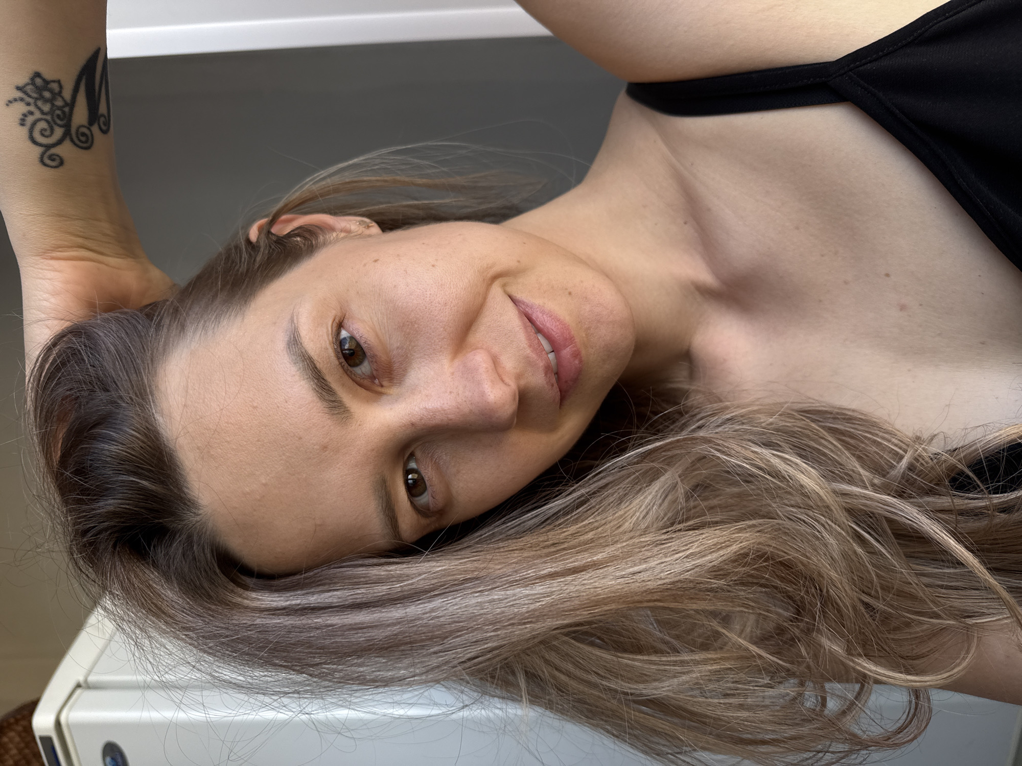 A close-up, eye-level portrait features a woman with long, light brown and blonde wavy hair, lying down and looking upwards towards the camera. Her head is slightly tilted, and her hair cascades over a light-colored surface beneath her. She has fair skin, a gentle, pleasant smile, and engaging brown eyes. Her lips are slightly parted, revealing a glimpse of her teeth. She wears a black top or camisole, with a strap visible over her shoulder. On her left wrist, a distinct black tattoo is visible, featuring a stylized letter "M" adorned with floral and swirl patterns. The background is simple and uniformly light, suggesting an indoor setting. The lighting is soft and natural, illuminating her face evenly. The overall mood is relaxed and friendly, capturing a casual moment in what is contextually identified as Chaikovsky, Russia.