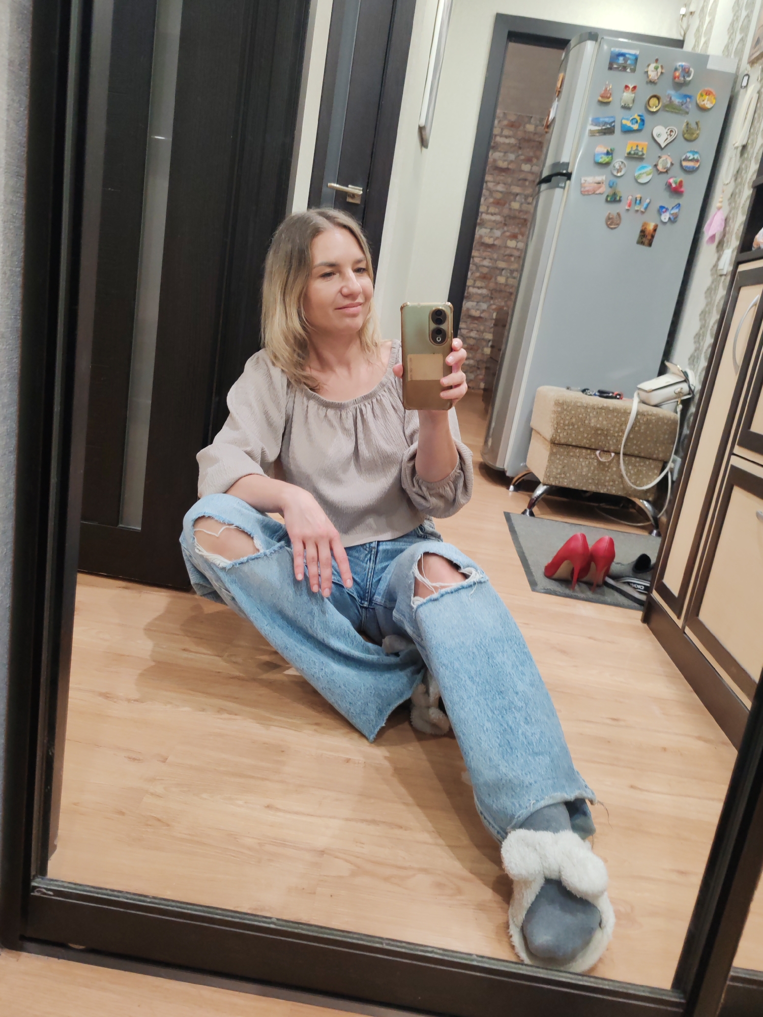 A woman is sitting cross-legged on a wooden floor in what appears to be a domestic interior. She's wearing a light gray blouse, ripped blue jeans, and gray slippers with white furry embellishments. She is holding a smartphone, appearing to take a selfie in a mirror.
In the background, there is a refrigerator covered with magnets and a cushioned ottoman. A pair of red high heels are visible near the ottoman. A kitchen cabinet is visible on the right.
Given the presence of what appears to be brick wallpaper, this image could be from any location, including Petropavlovsk-Kamchatsky, Russia, but there are no definitive visual cues to confirm that location.