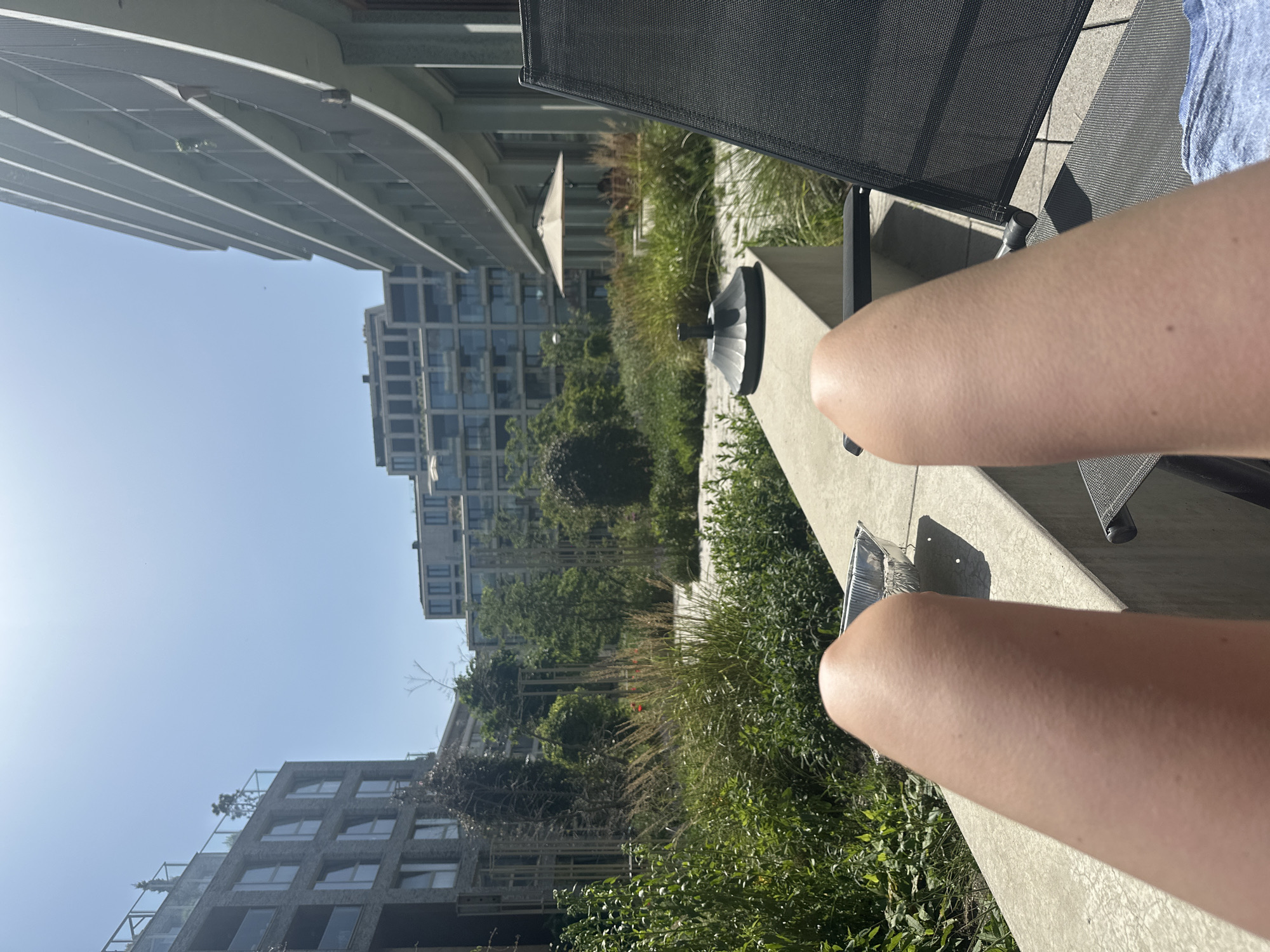 The image shows a person sitting on a chair on a balcony overlooking a residential area in Amsterdam, Netherlands. The person's legs are visible in the foreground, with a black mesh chair and a concrete ledge. In the background, there are several modern apartment buildings interspersed with trees and green spaces. The sky is blue, and the overall scene suggests a sunny day. The location is likely a higher floor of one of the residential buildings, providing a view of the surrounding neighborhood. An aluminum container sits on the ledge.