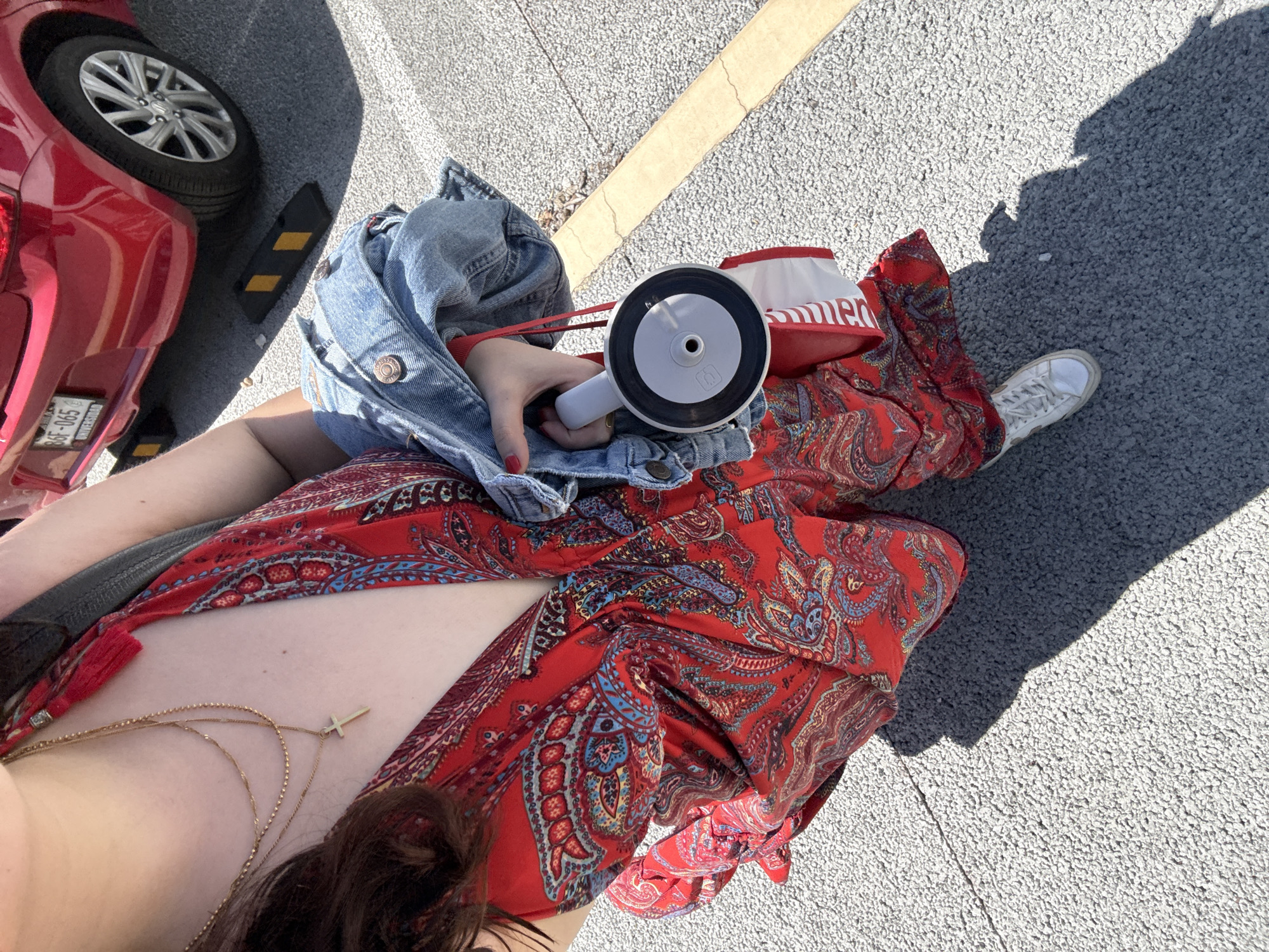 An overhead shot captures a person and their surroundings in what appears to be an outdoor parking area in bright daylight. The person, mostly visible from the torso down, is wearing a flowing red dress with an intricate paisley pattern in shades of blue, red, and gold. A denim jacket is casually draped over their lap or arm. A delicate gold necklace with a cross pendant is visible against their chest. They are holding a white insulated bottle with a black ring around its top and a small, indistinct square logo. Next to the bottle, a red and white item, possibly a sun hat or bag, displays the word "Sommer" in white letters. The person is also wearing white sneakers with beige soles.
To the left, a red car is partially visible, showing its rear wheel, a portion of the bumper, and a black and yellow concrete parking barrier. The car's license plate clearly displays "36E-065". The ground is covered in light gray, coarse gravel, with a faint yellow parking line running horizontally across the upper right. A large, dark shadow from an unseen object or person is cast prominently on the gravel in the mid-right portion of the image. The strong shadows suggest it is a sunny day.