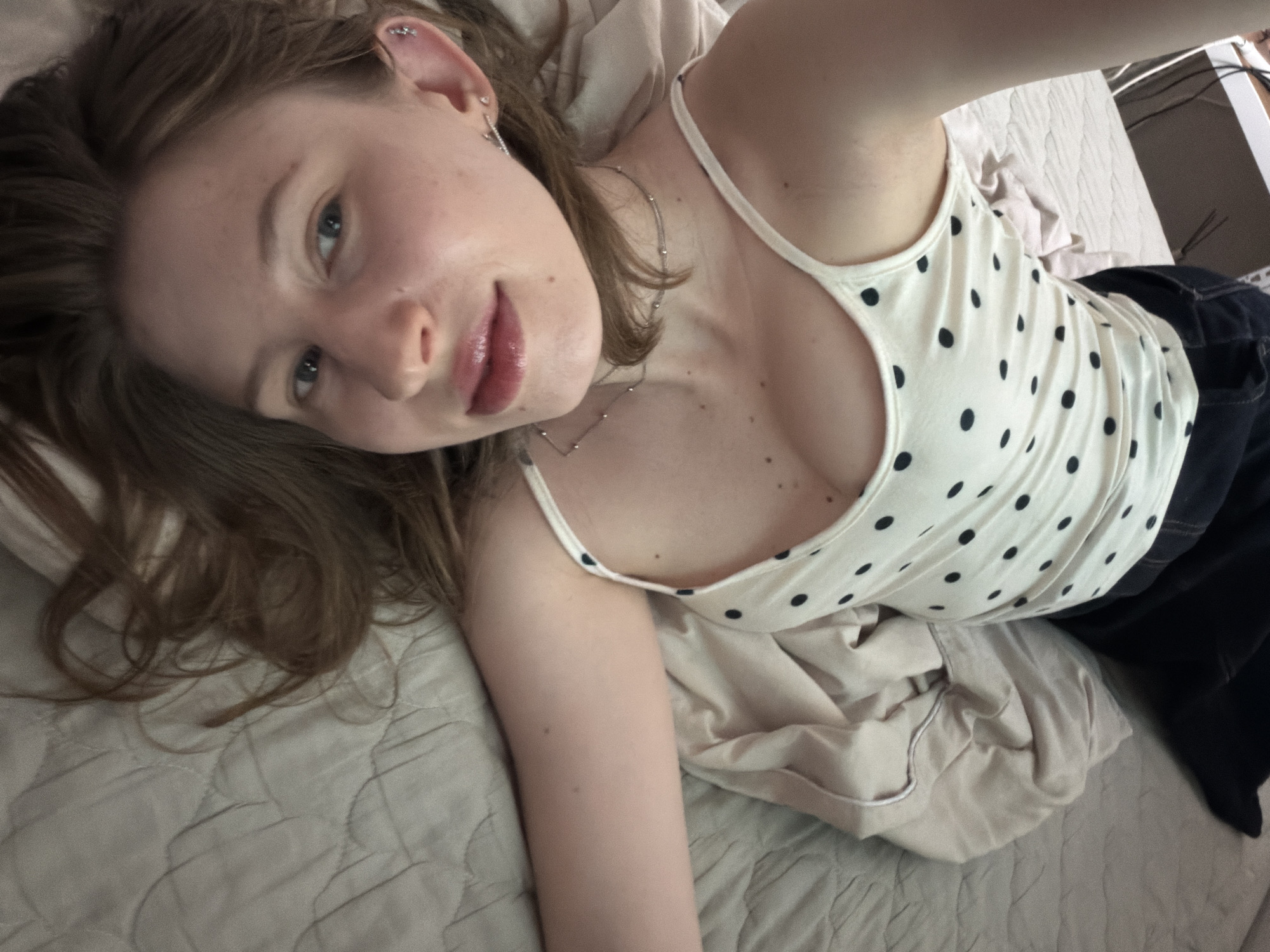 The image shows a young woman lying on a bed, facing upwards. She has long, brown hair that is slightly messy and frames her face. Her eyes are a light blue and she is looking directly at the camera. She is wearing a white tank top with black polka dots and a thin silver necklace. Her lips are painted with a glossy pink lipstick. Her left ear has multiple piercings with small silver earrings. The background is a bed with light-colored bedding that appears to be quilted. The lighting suggests it is daytime, possibly indoors. The overall mood of the image is relaxed and intimate. There is no visible text in the image. The location context of Dongducheon-si, South Korea, does not provide any specific visual cues in this particular image.
