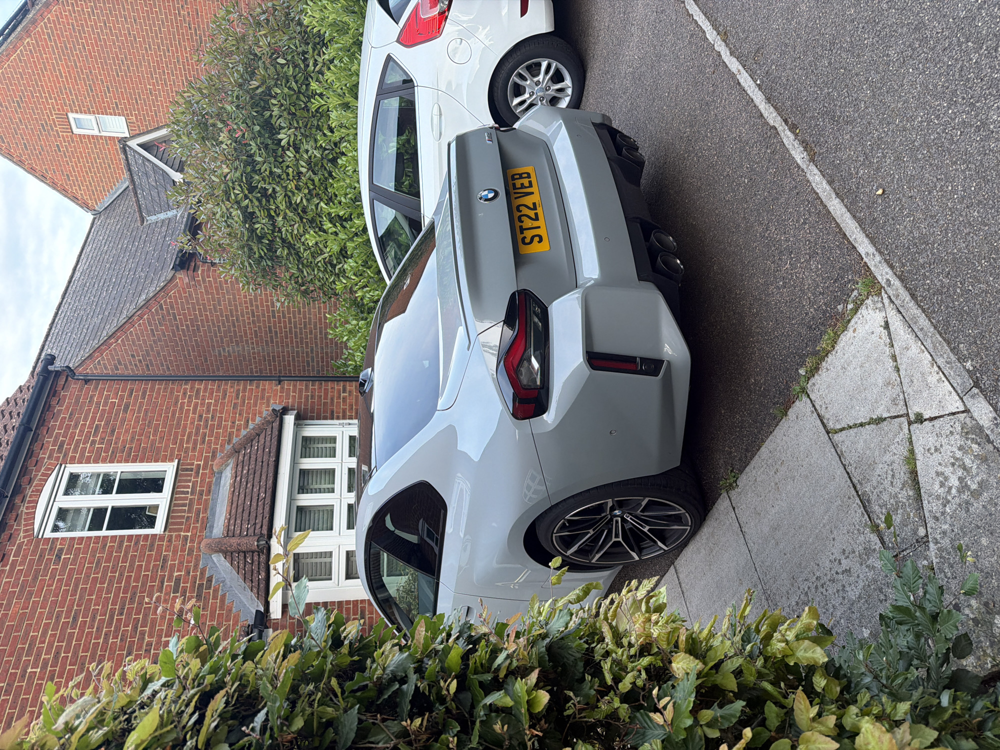 This image depicts a static daytime scene on a residential street in Maidstone, United Kingdom, characterized by an overcast sky. The central subject is a light grey BMW M2 coupe (G87 generation), registered with the UK license plate "ST22 VEB" (indicating a 2022 registration). The vehicle is positioned on grey concrete paving slabs, partially obscured on the left by a dense green hedge exhibiting some yellowish leaves. The BMW features black multi-spoke alloy wheels, a black roof, a body-colored rear spoiler lip, and a prominent rear diffuser accommodating four circular exhaust outlets.
Behind the BMW, a white, five-door Ford Fiesta is partially visible, parked on a dark asphalt surface that shows a faded white line along its edge.
To the left of the frame, a red brick house features a white-framed bay window on the ground floor (partially obscured) and a white-framed, arched-top window on the first floor. This house has a brown tiled roof with visible moss accumulation. A black rainwater downpipe is visible on its right side. In the background, a second red brick house is partially visible, displaying a small, high-set rectangular window and a darker, possibly slate-tiled roof. A large green shrub with some reddish foliage stands between the two houses.
No individuals are present. The vehicles are stationary, and the overall scene conveys a typical, quiet residential environment. Weeds are observed growing in the cracks of the concrete paving and along the edge of the asphalt.