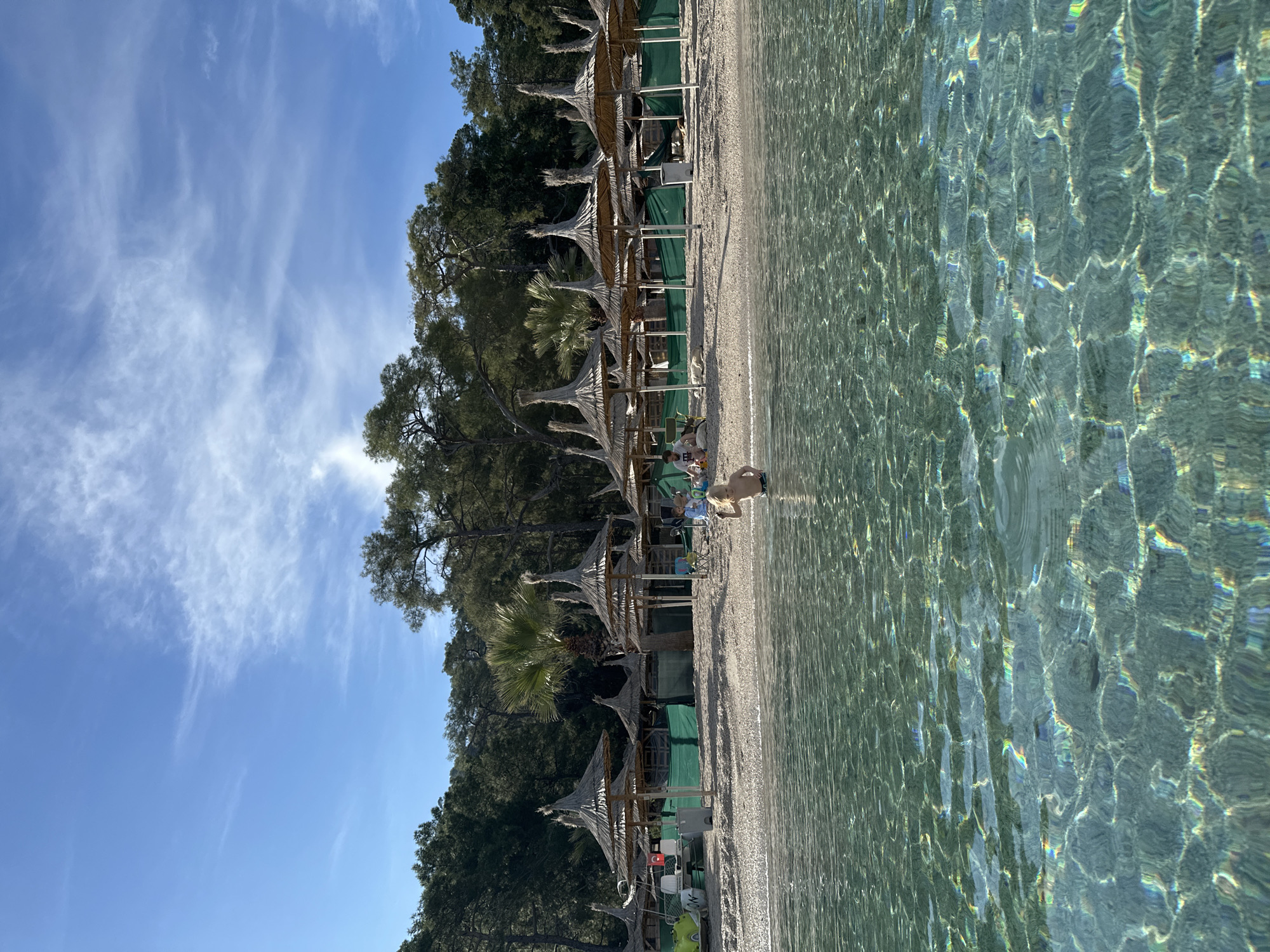 The image depicts a beach scene in Unknown, Türkiye. The scene is oriented with the beach running vertically. Along the left side of the image is a sandy beach lined with thatched umbrellas and beach chairs. Trees and vegetation border the beach, reaching the sky.
In the center-right of the image, a boy stands at the edge of the water, partially submerged. The water is clear and reflects sunlight, creating patterns of light and shadow.
Above the trees, a blue sky is visible with wispy clouds. The overall impression is of a sunny day at a beach resort.